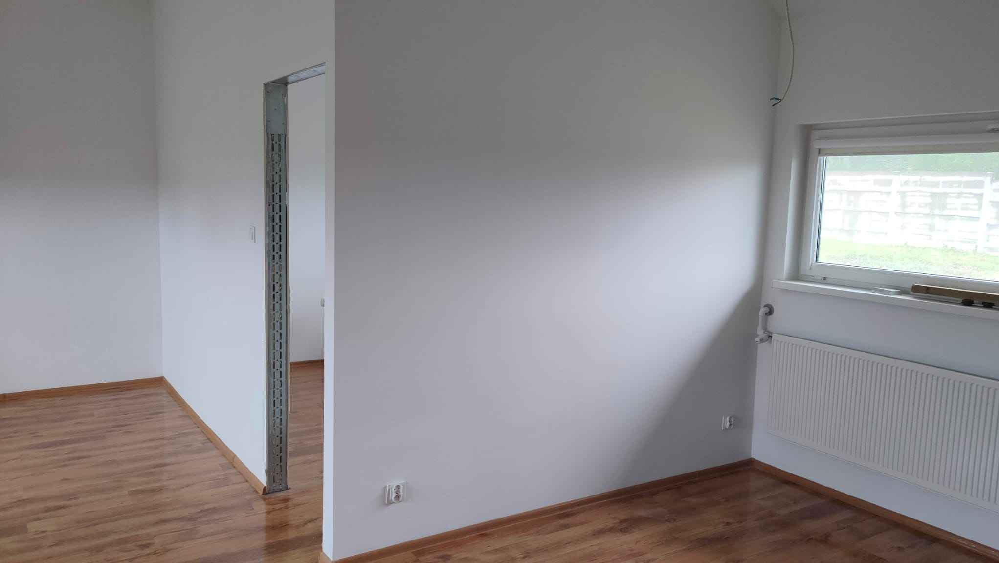 Pomalowane na biało wnętrze pokoju z drewnianą podłogą, grzejnikiem pod oknem i metalową futryną drzwiową bez drzwi.