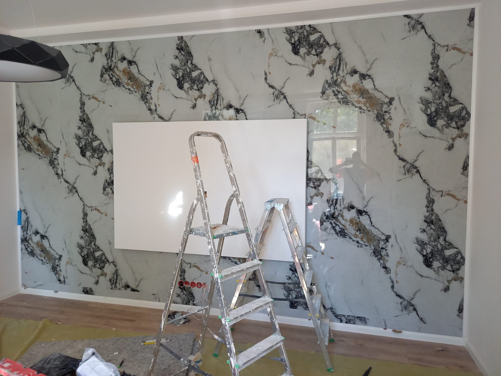 Ściana wykończona płytami imitującymi marmur z białym panelem, dwie drabiny malarskie, widoczne gniazdka elektryczne. Prace wykończeniowe w trakcie realizacji.