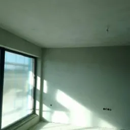 Wnętrze nowo wykończonego pokoju z dużym oknem, ściany i sufit pokryte świeżym, jasnoszarym tynkiem, widoczne ślady po gniazdkach elektrycznych, okno zabezpieczone folią ochronną.