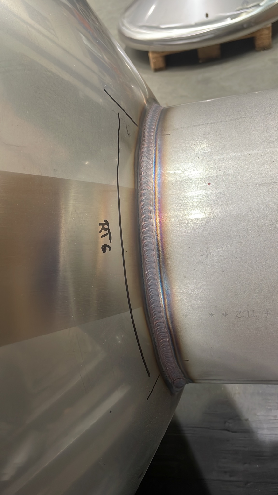 Połączenie spawane dwóch elementów metalowych, widoczne ślady po spawaniu, czarne oznaczenia 'RT6' i linia pomocnicza obok spoiny.