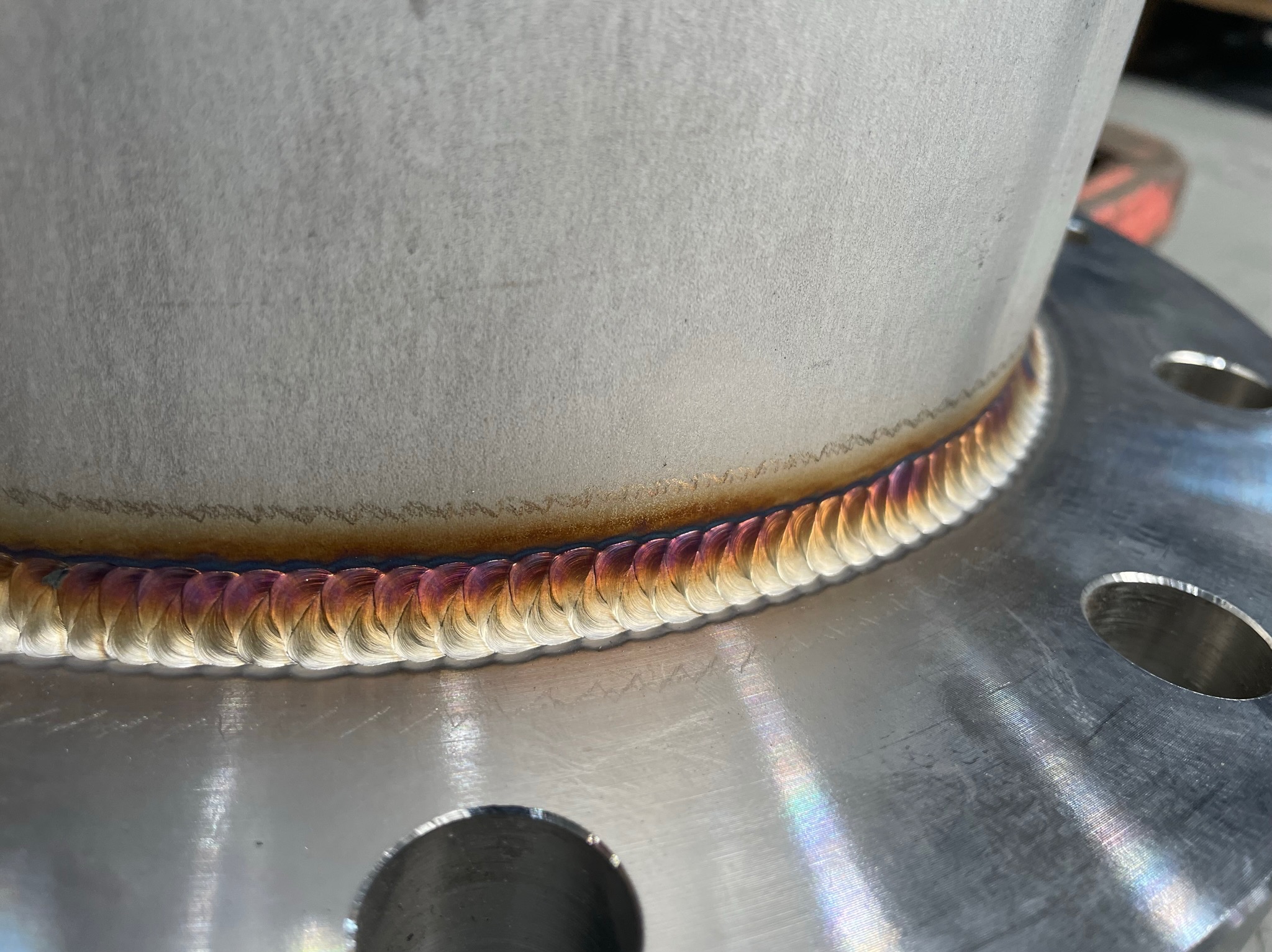 Precyzyjne spoiny na metalowej rurze z kołnierzem, widoczne tęczowe przebarwienia termiczne.