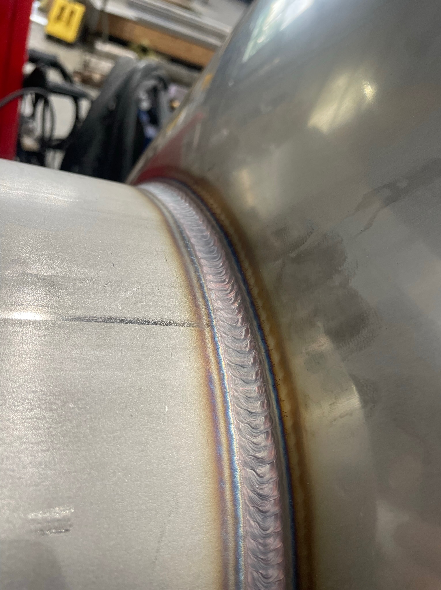 Szczegółowe zbliżenie na spaw dwóch metalowych rur, widoczne ślady spawania z opalizującymi kolorami, tło warsztatowe.