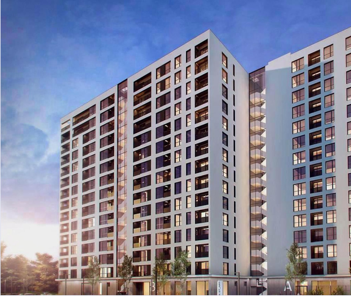 Wysoki budynek mieszkalny z jasną elewacją i licznymi oknami, widoczny z dołu pod kątem, z przeszklonym łącznikiem między segmentami i drzewami w tle.