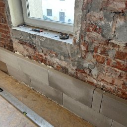 TOPAZ YEHOR TOPALOV - Wyrównywanie ściany z cegły bloczkami z betonu komórkowego pod oknem. Widoczne narzędzia: poziomica, śrubokręt. Etap prac remontowych w pomieszczeniu.