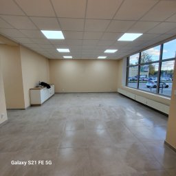 TOPAZ YEHOR TOPALOV - Przestronne, jasne wnętrze po remoncie z beżowymi ścianami, szarą podłogą z płytek i dużym oknem. Widoczne grzejniki i szafka przy ścianie. Sufit z kwadratowymi lampami.