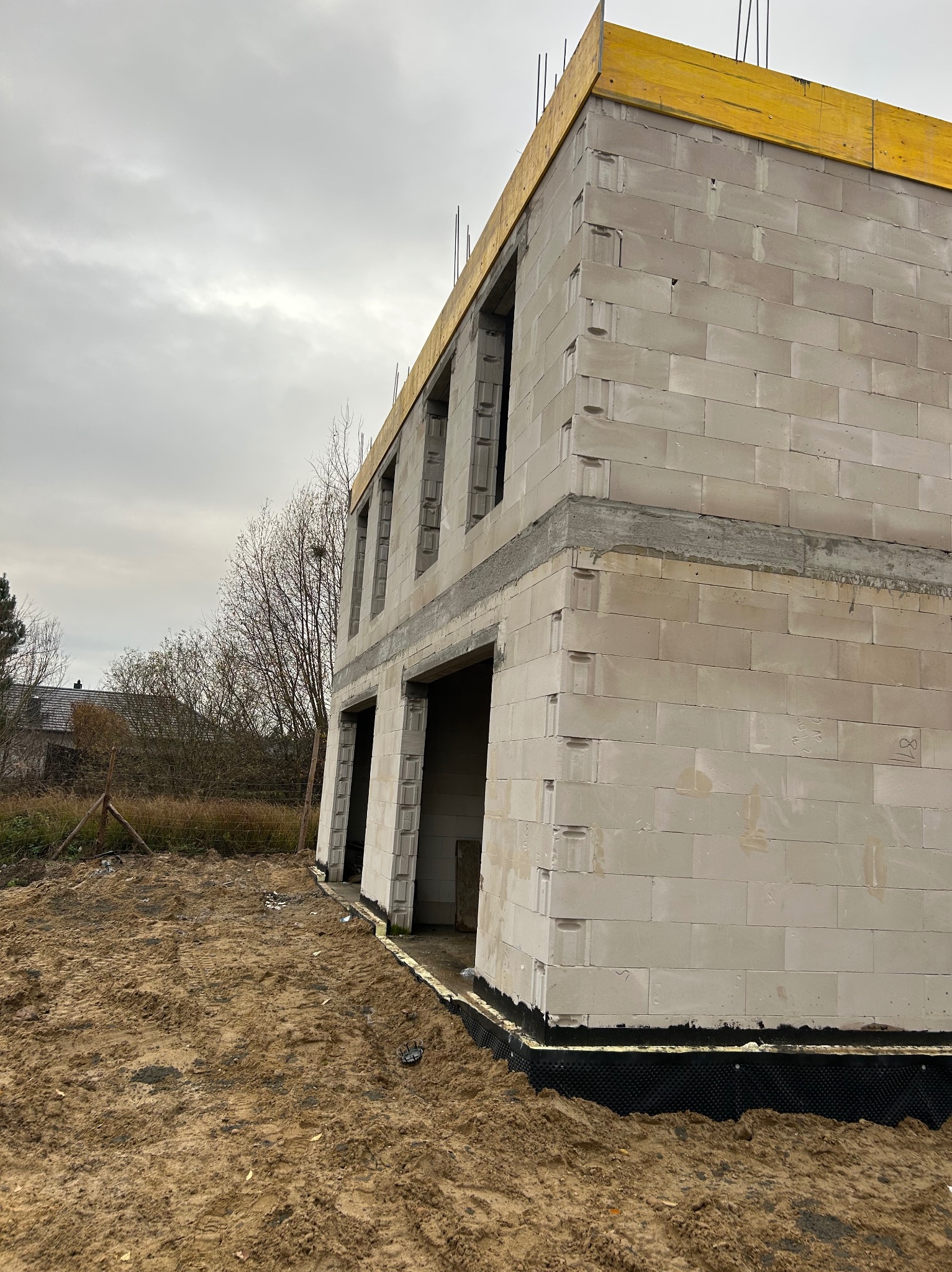 Surowy stan dwukondygnacyjnego domu z betonu komórkowego, widoczne otwory okienne i drzwiowe, żółte deski szalunkowe na górze, grunt piaszczysty.