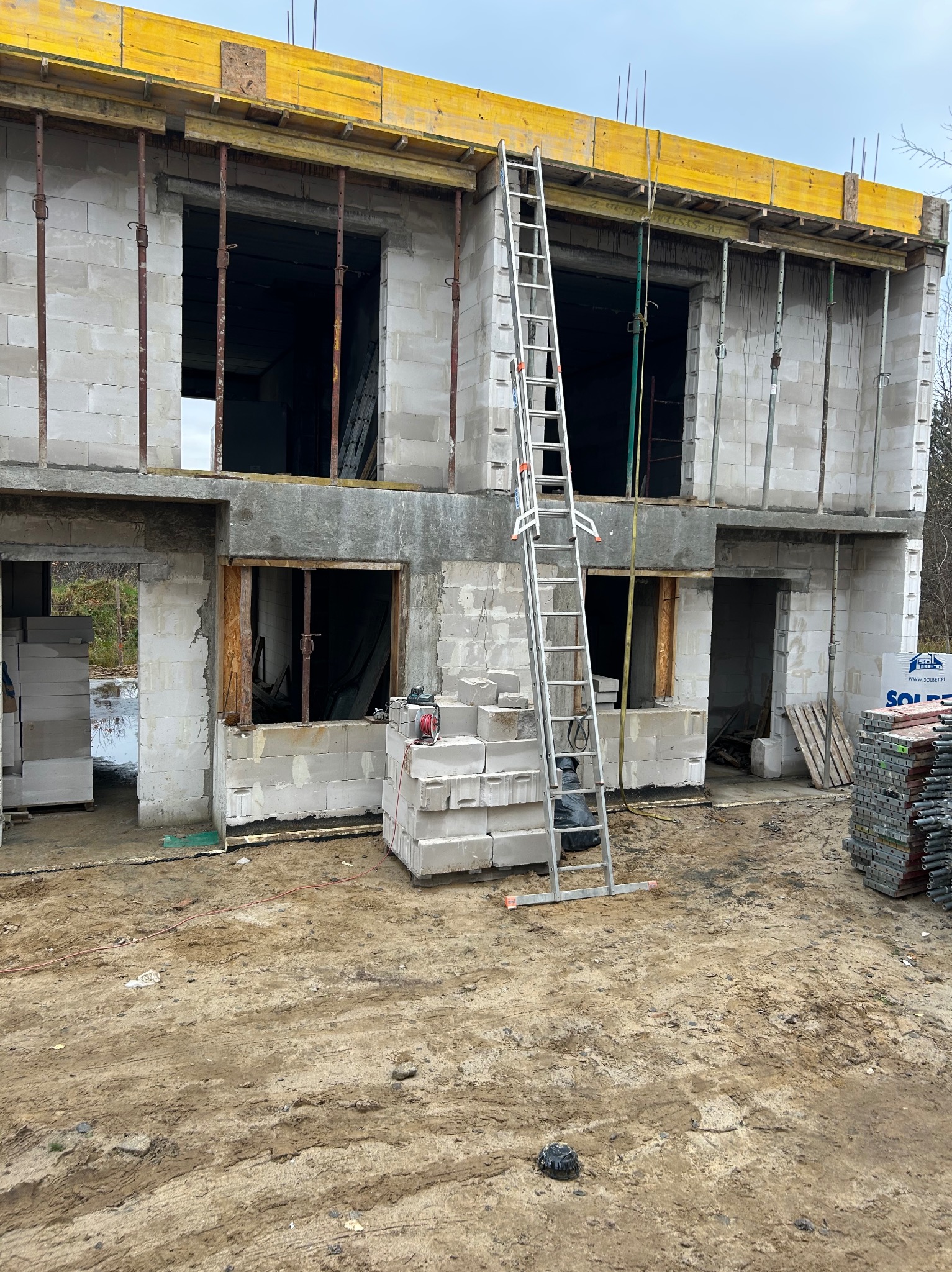 Surowy stan domu z białego pustaka, widoczne szalunki i podpory stropu, drabina oparta o ścianę. Etap budowy z widocznymi materiałami budowlanymi.