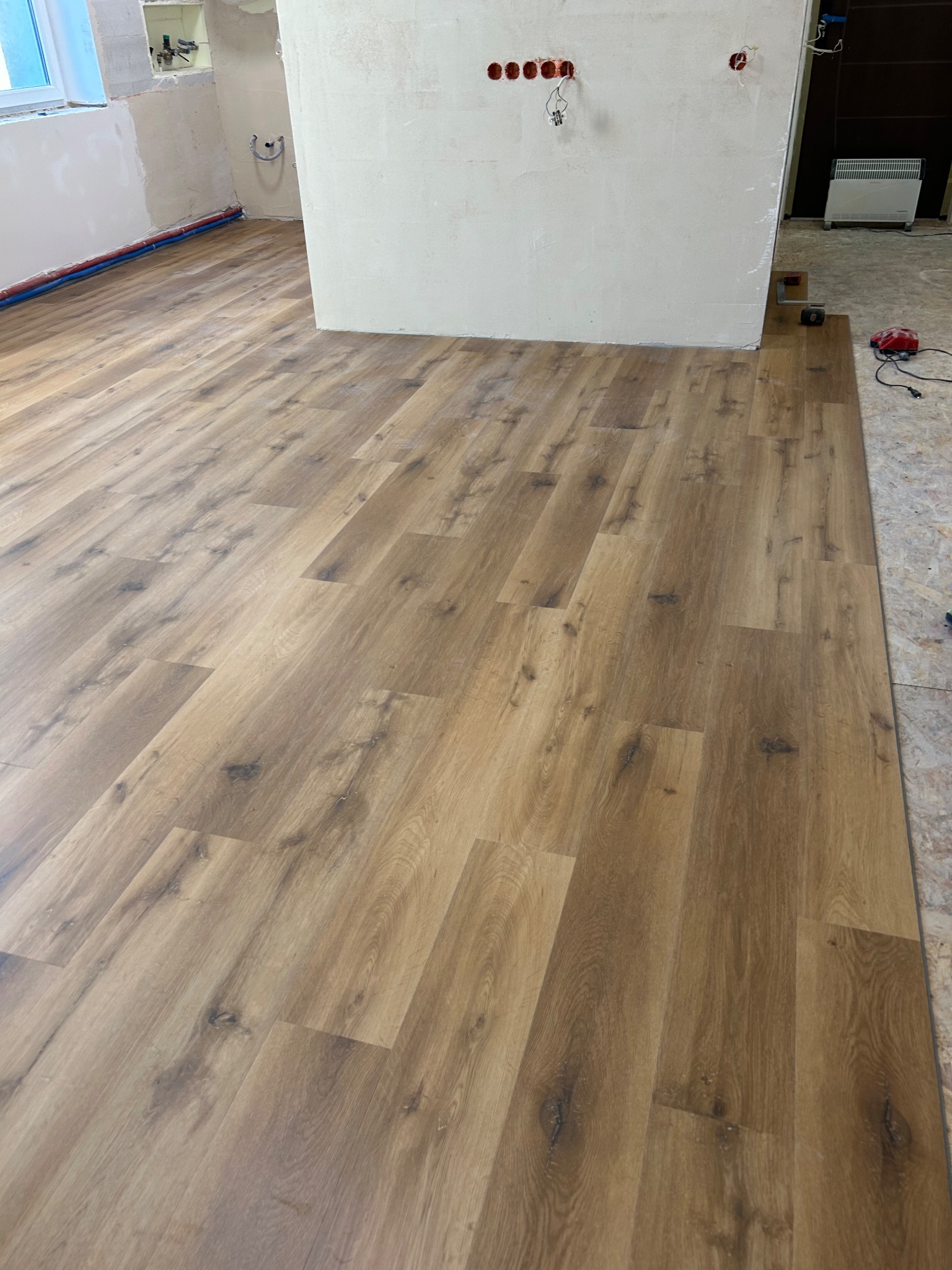 Układanie paneli podłogowych w jasnym odcieniu drewna w trakcie remontu. Widoczne niedokończone ściany i wystające kable. Częściowo ułożona podłoga.