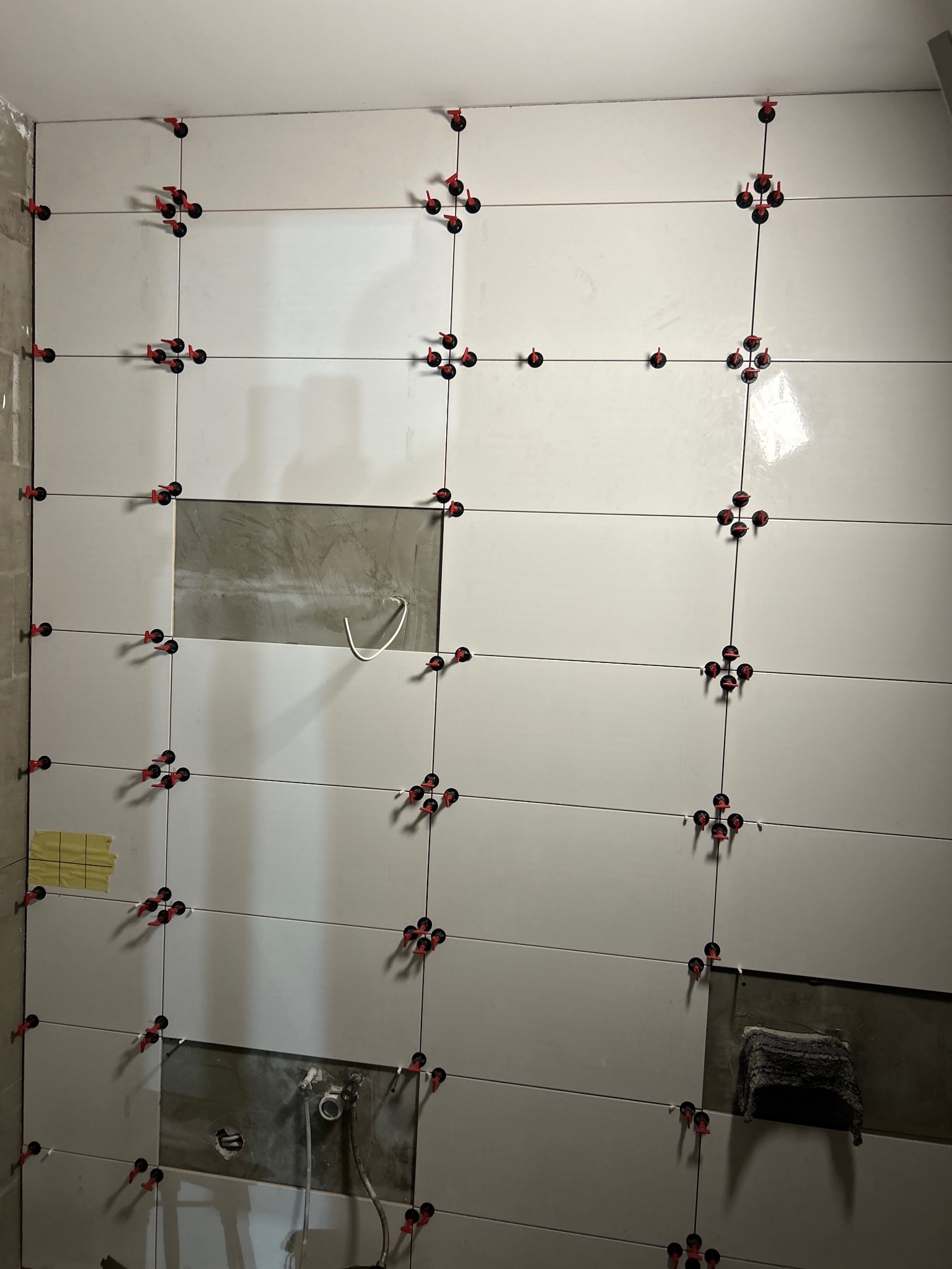 Ściana z białych, błyszczących płytek w trakcie układania, z widocznymi systemami poziomowania (czerwone klipsy i czarne kliny). Fragmenty ściany bez płytek, widoczne przygotowane podłoże.