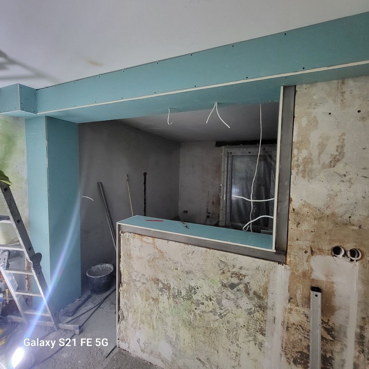 Wnętrze w trakcie remontu: ściany z surowym tynkiem, częściowo obłożone płytami gipsowo-kartonowymi w kolorze turkusowym, widoczne przewody elektryczne i drabina.