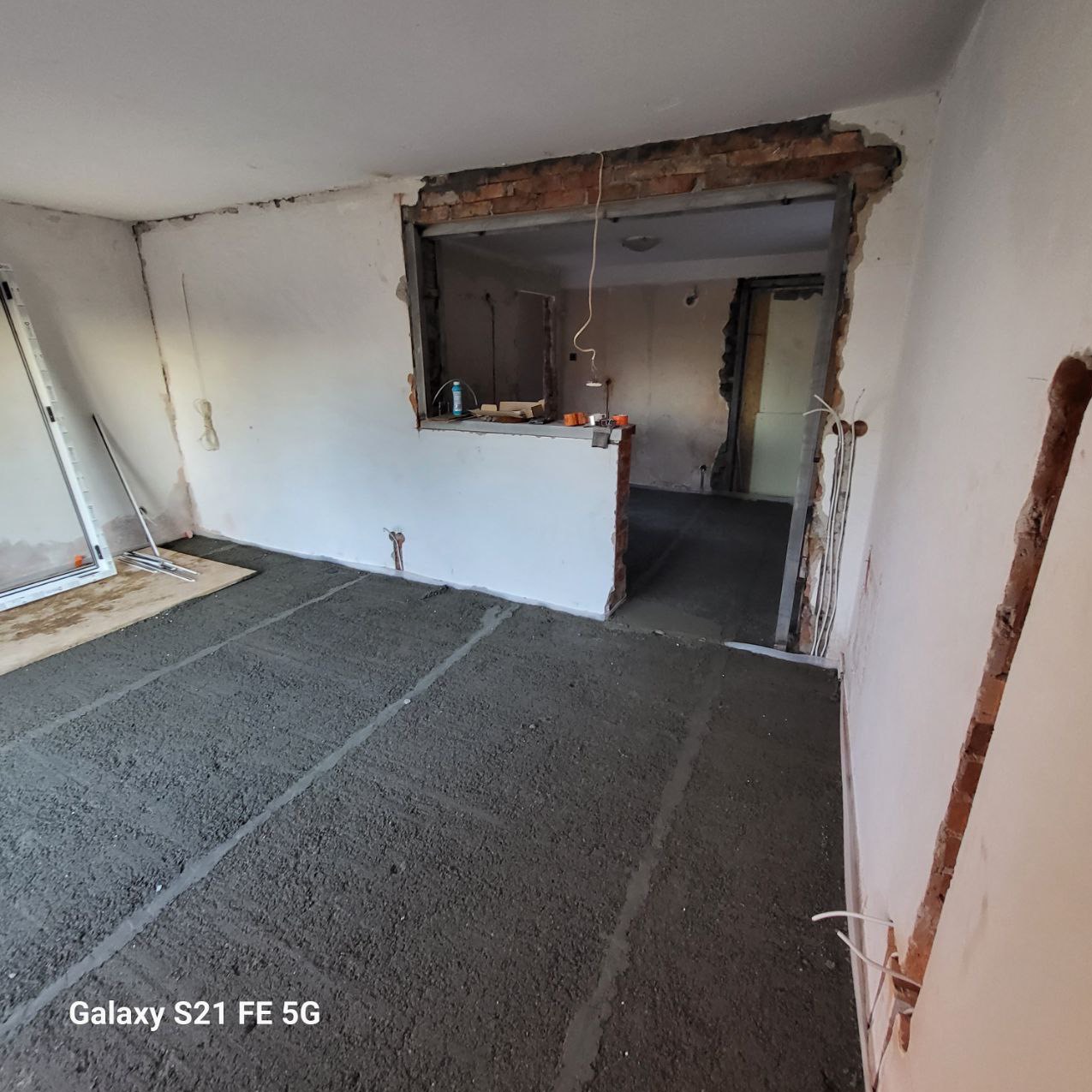 Wnętrze w trakcie remontu: wylana posadzka cementowa z widocznymi śladami po łatach, ściany przygotowane do szpachlowania, otwór w ścianie z odsłoniętą cegłą.