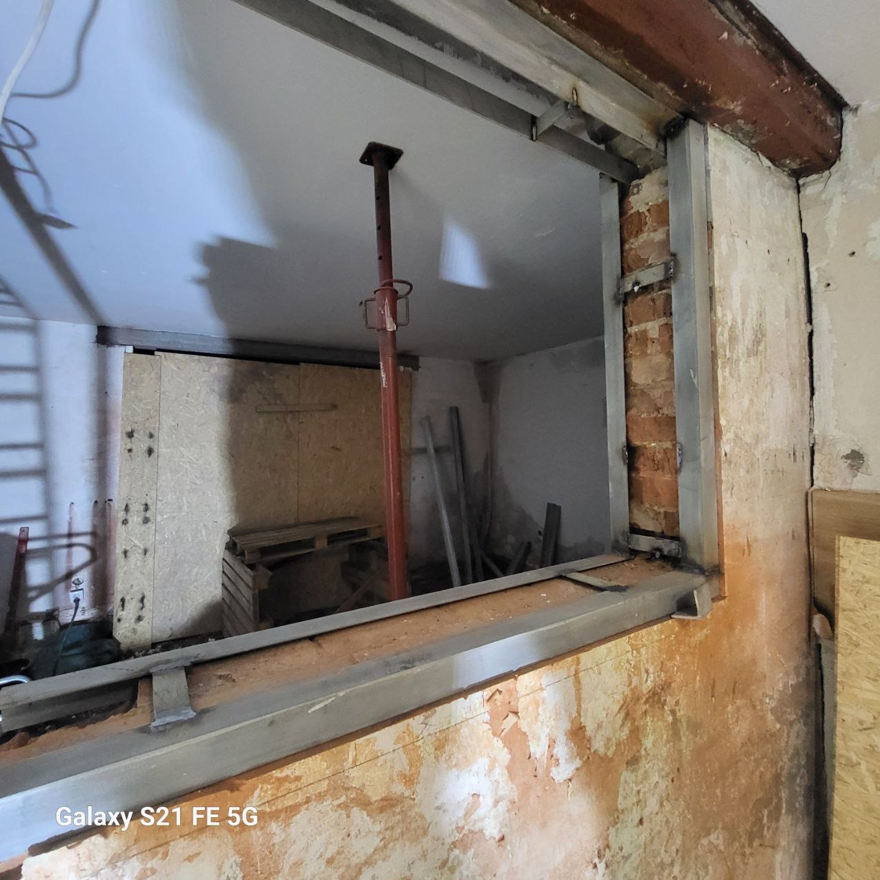 Wzmocnienie otworu okiennego stalową konstrukcją w trakcie remontu. Widoczne cegły, wsporniki i surowy stan ścian. Prace remontowe w pomieszczeniu.