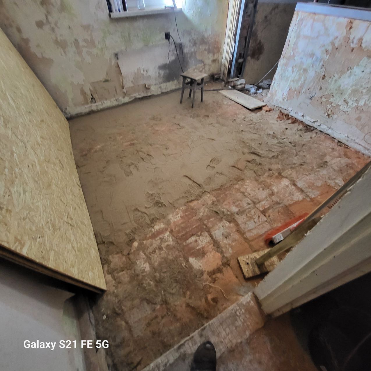 Pokój w trakcie remontu: ściany z resztkami farby, podłoga z cegieł pokryta warstwą zaprawy, płyta OSB oparta o ścianę. Widoczne ślady stóp w zaprawie.