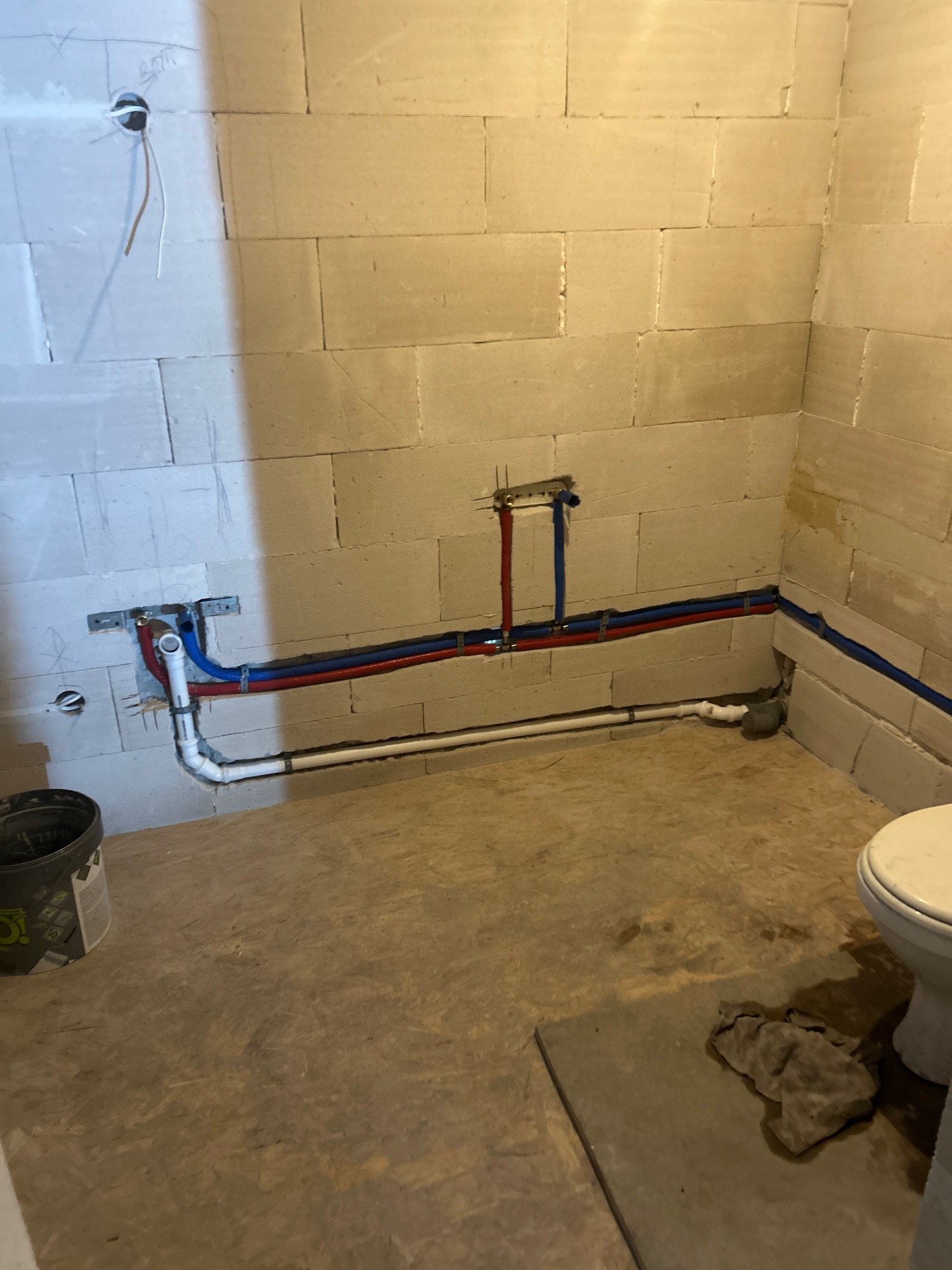 Surowa łazienka w trakcie instalacji wodno-kanalizacyjnej: widoczne rury (czerwone, niebieskie, białe) zamocowane na ścianie z bloczków, przygotowane podłączenie toalety.