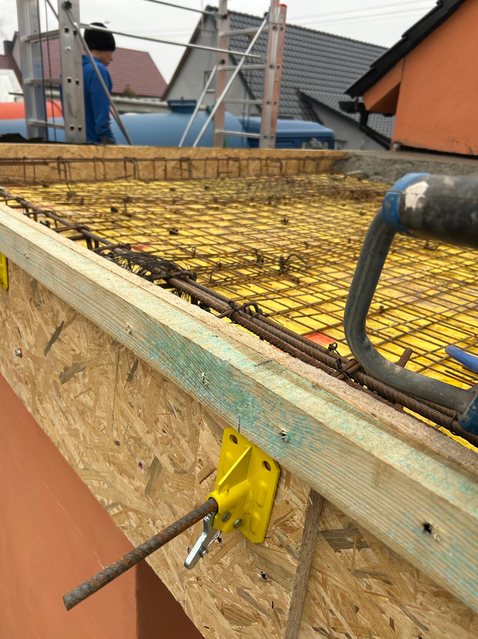 Zbrojenie stropu z żółtymi elementami mocującymi i siatką na żółtej folii. W tle widoczny pracownik na dachu i zabudowa jednorodzinna.