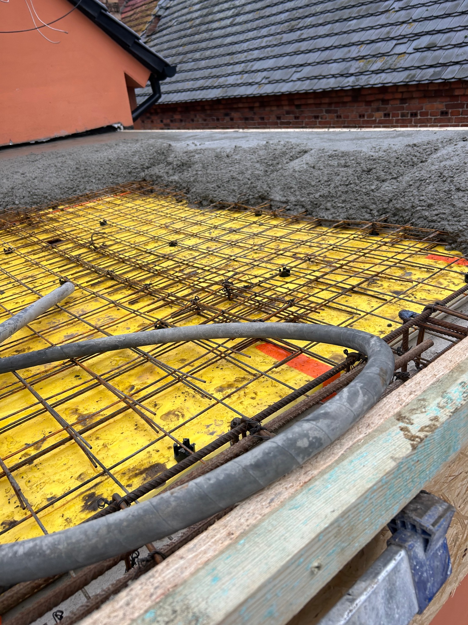 Zbrojenie stropu z siatki stalowej na żółtej płycie szalunkowej, częściowo zalane betonem. Widoczne elementy konstrukcyjne i przewody. Ujęcie z bliska, z góry.