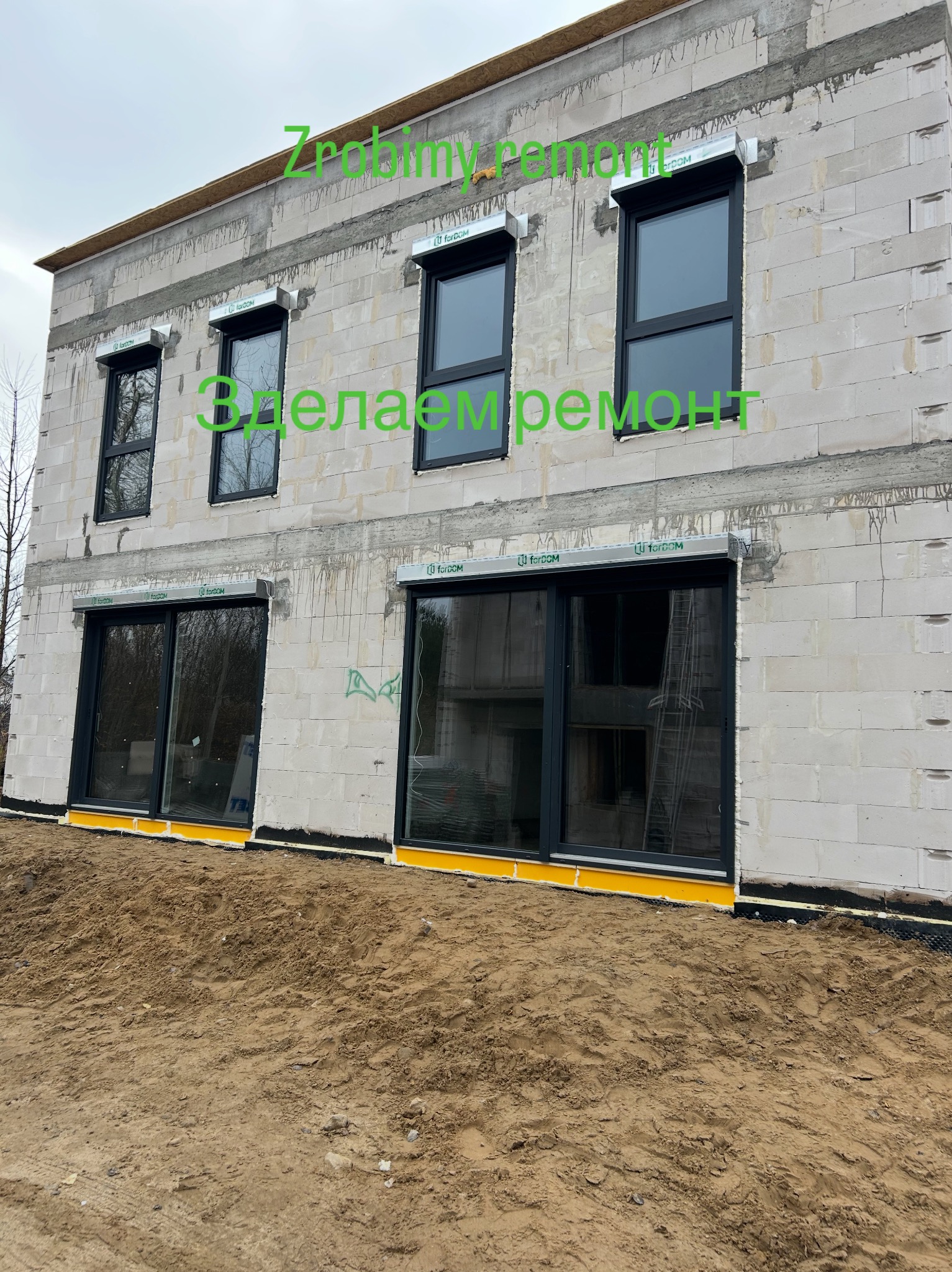 Surowy stan domu z betonu komórkowego z zamontowanymi oknami i roletami zewnętrznymi. Widoczne żółte zabezpieczenia okien. Na ścianie napisy w obcym języku.