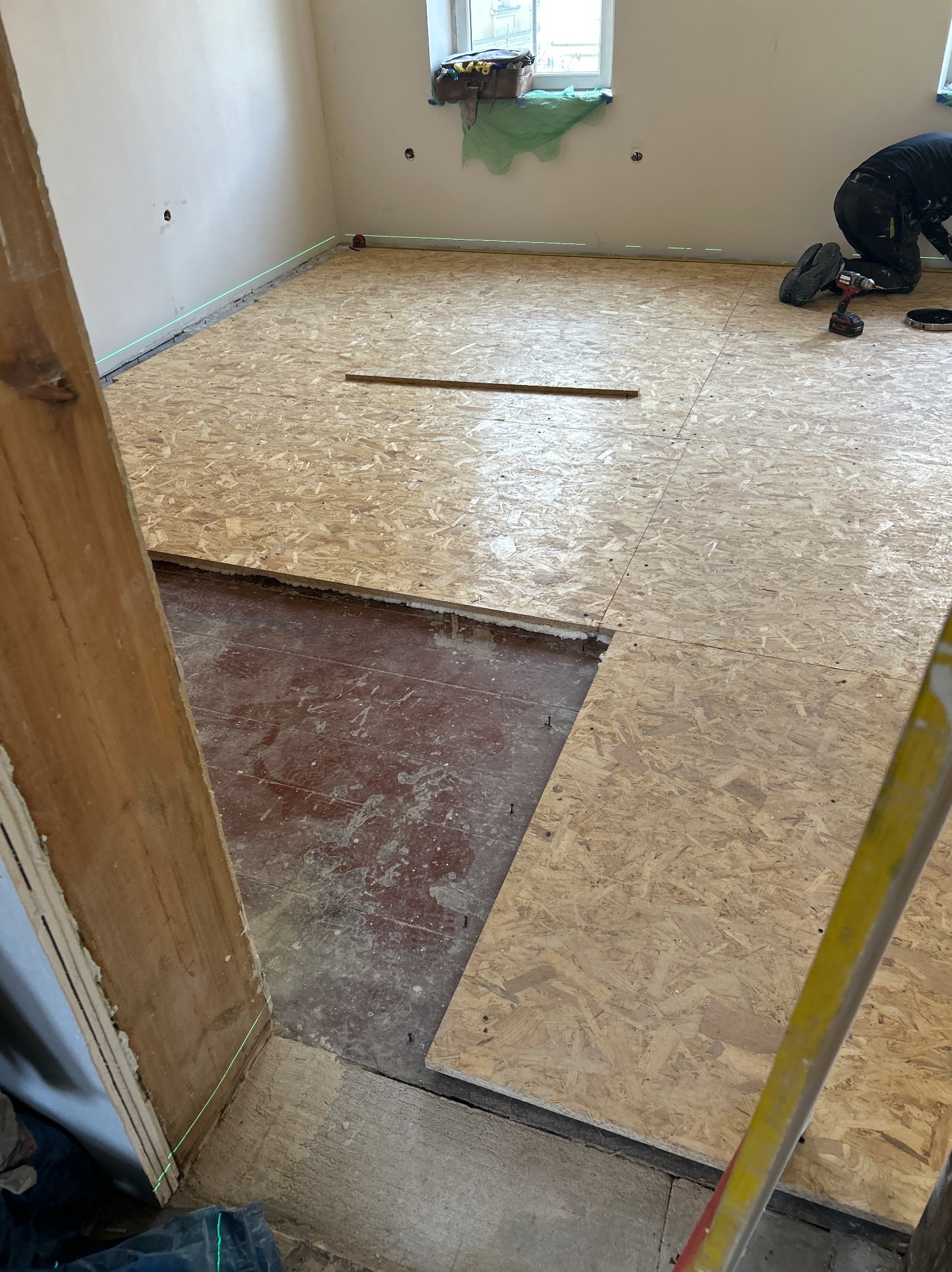 Układanie płyt OSB na podłodze w pokoju, widoczne narzędzia i laserowy niwelator. Część podłogi jeszcze nie pokryta płytami, widoczna stara podłoga.