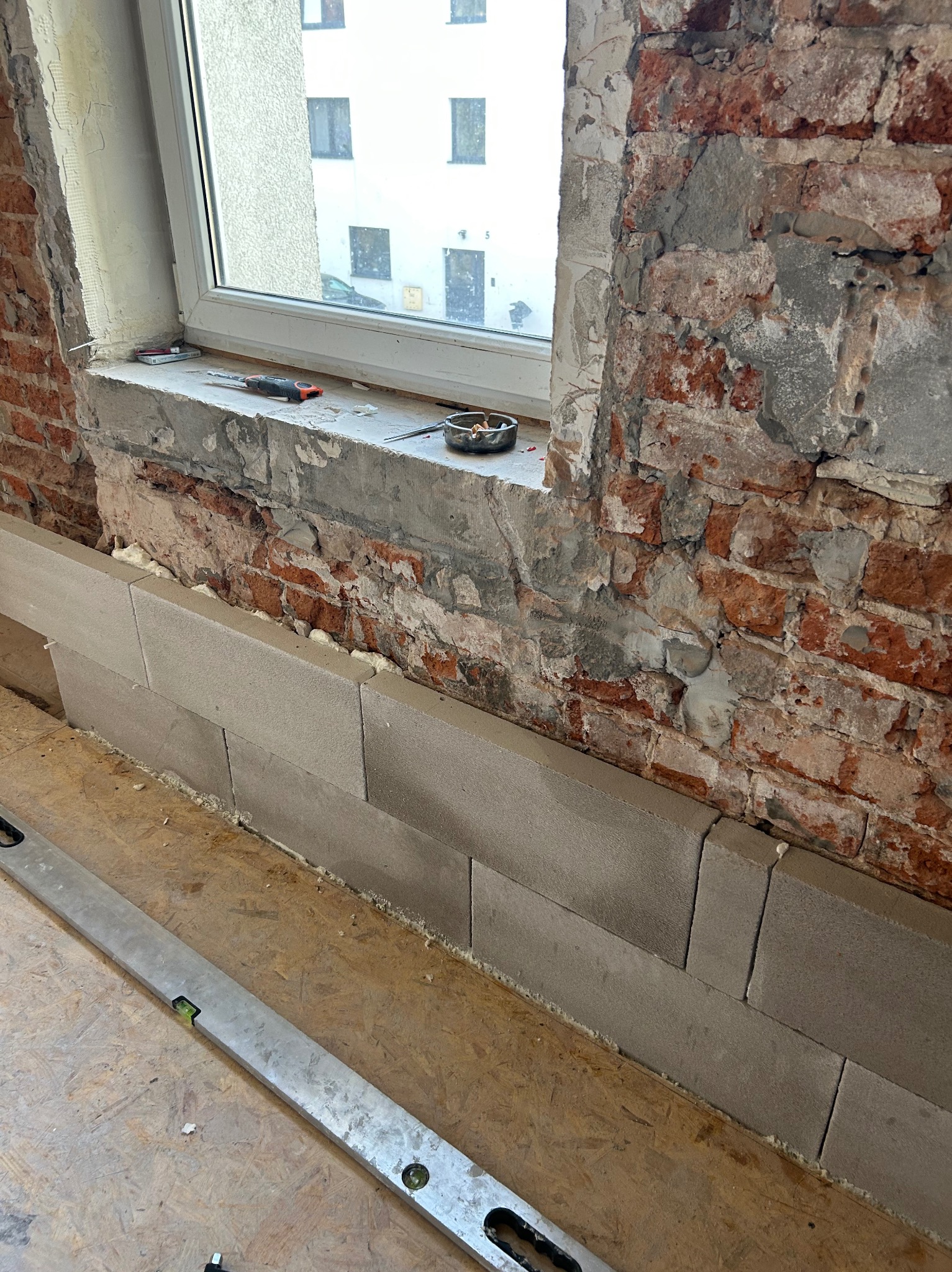 Wyrównywanie ściany z cegły bloczkami z betonu komórkowego pod oknem. Widoczne narzędzia: poziomica, śrubokręt. Etap prac remontowych w pomieszczeniu.