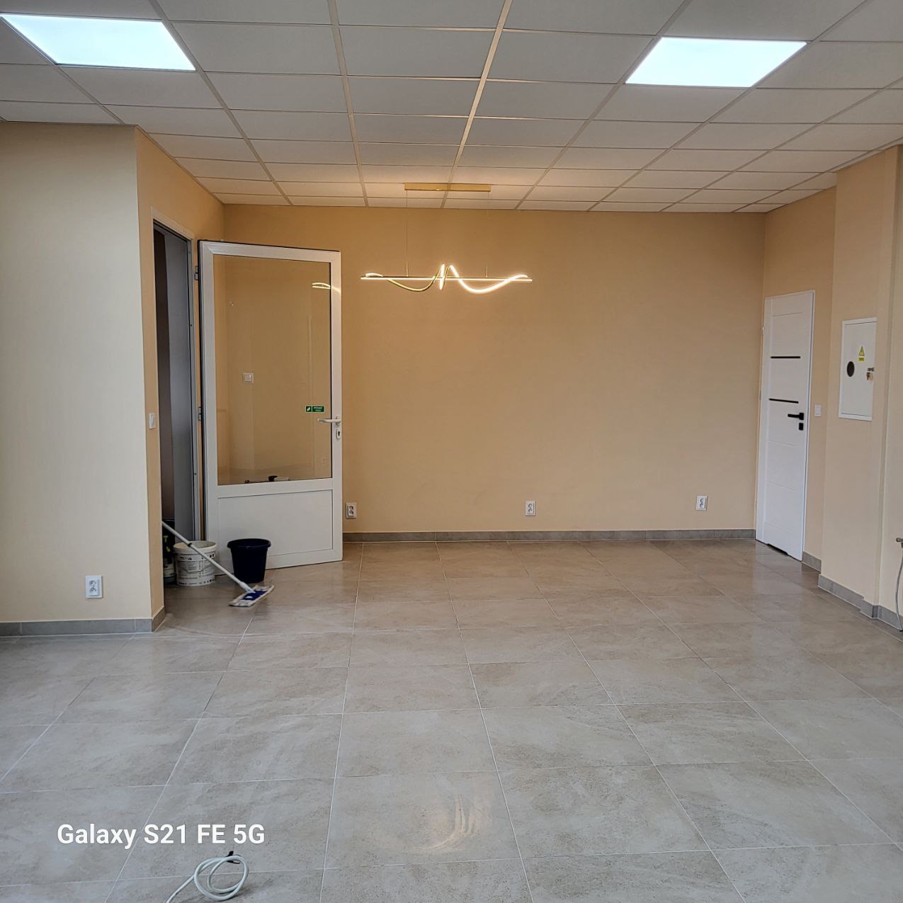 Jasne wnętrze po remoncie: beżowe ściany, szara podłoga z płytek, nowoczesna lampa. Widoczne wiadro i mop sugerują świeże sprzątanie po pracach wykończeniowych.