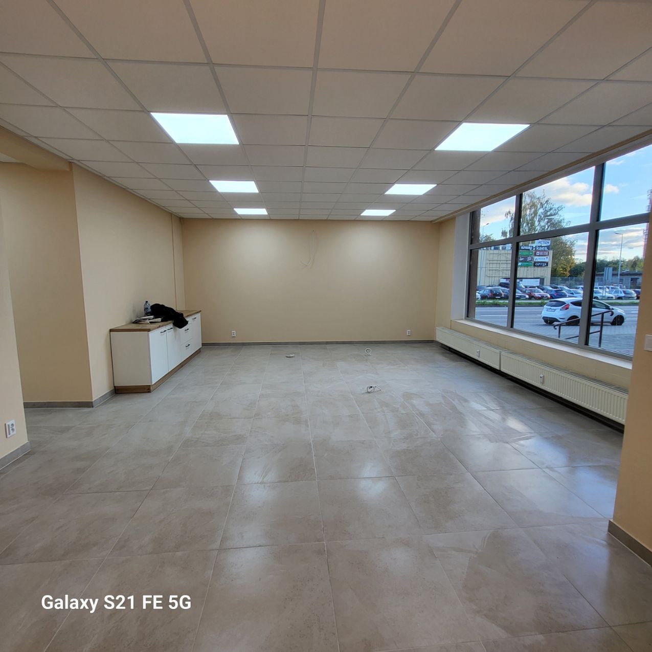 Przestronne, jasne wnętrze po remoncie z beżowymi ścianami, szarą podłogą z płytek i dużym oknem. Widoczne grzejniki i szafka przy ścianie. Sufit z kwadratowymi lampami.