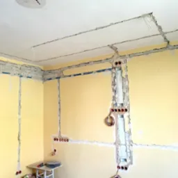 Żółta ściana w trakcie montażu instalacji elektrycznej, widoczne kable w bruzdach i puszki pod gniazdka, biały sufit z wyżłobionymi kanałami na przewody, mały stolik z narzędziami w rogu pokoju.