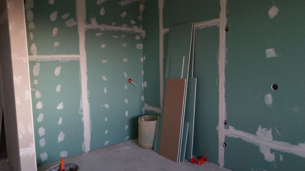 Wnętrze w trakcie wykańczania: ściany z płyt gipsowo-kartonowych w kolorze zielonym, z widocznymi łączeniami szpachlowanymi na biało, przewody elektryczne wystające ze ściany, płyty gipsowe oparte...