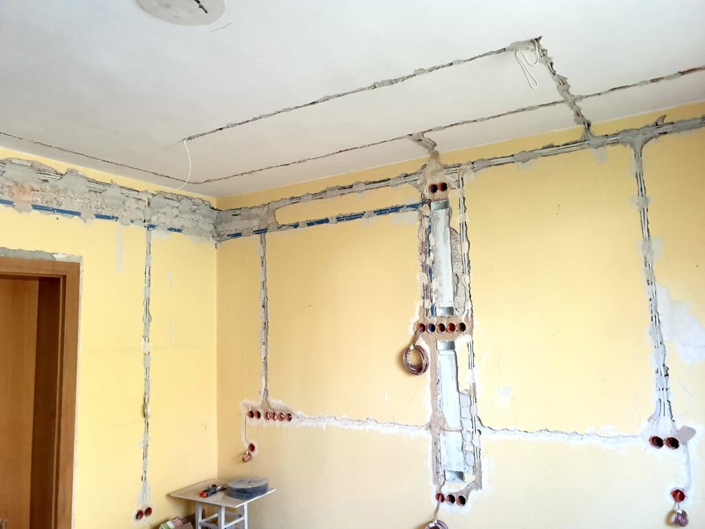 Żółta ściana w trakcie montażu instalacji elektrycznej, widoczne kable w bruzdach i puszki pod gniazdka, biały sufit z wyżłobionymi kanałami na przewody, mały stolik z narzędziami w rogu pokoju.