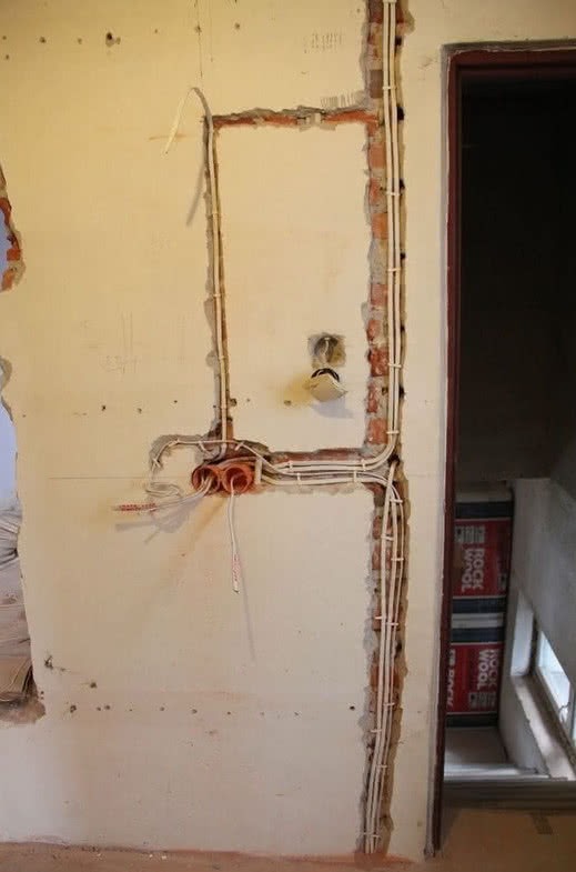 Wewnętrzna instalacja elektryczna w trakcie montażu: widoczne kable w białych rurkach PCV prowadzone w bruzdach w ścianie, puszki elektryczne i wystające przewody. Ściana w stanie surowym,...