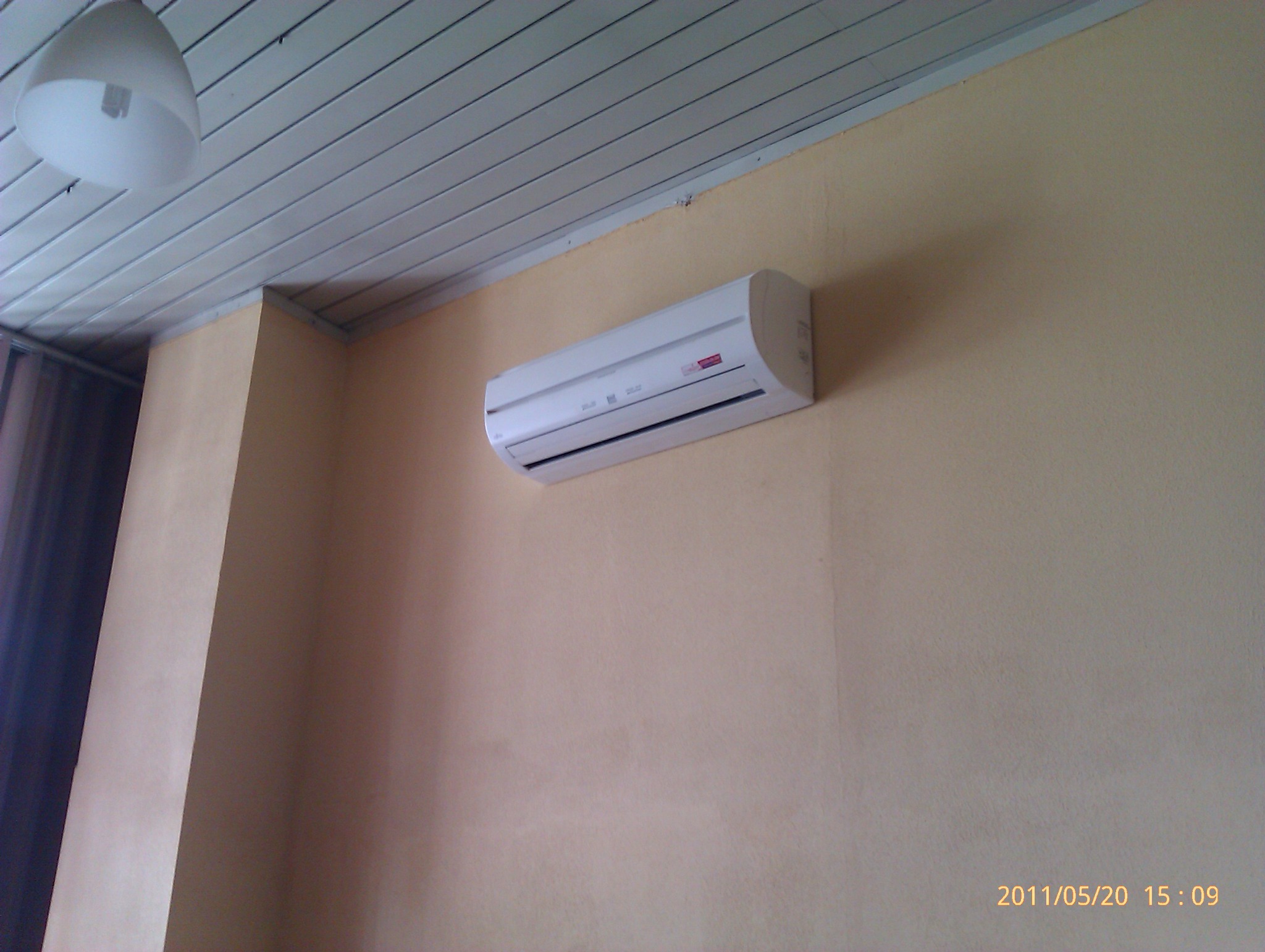 Biały klimatyzator ścienny zamontowany na beżowej ścianie, widoczny fragment sufitu z lampą i listwami.