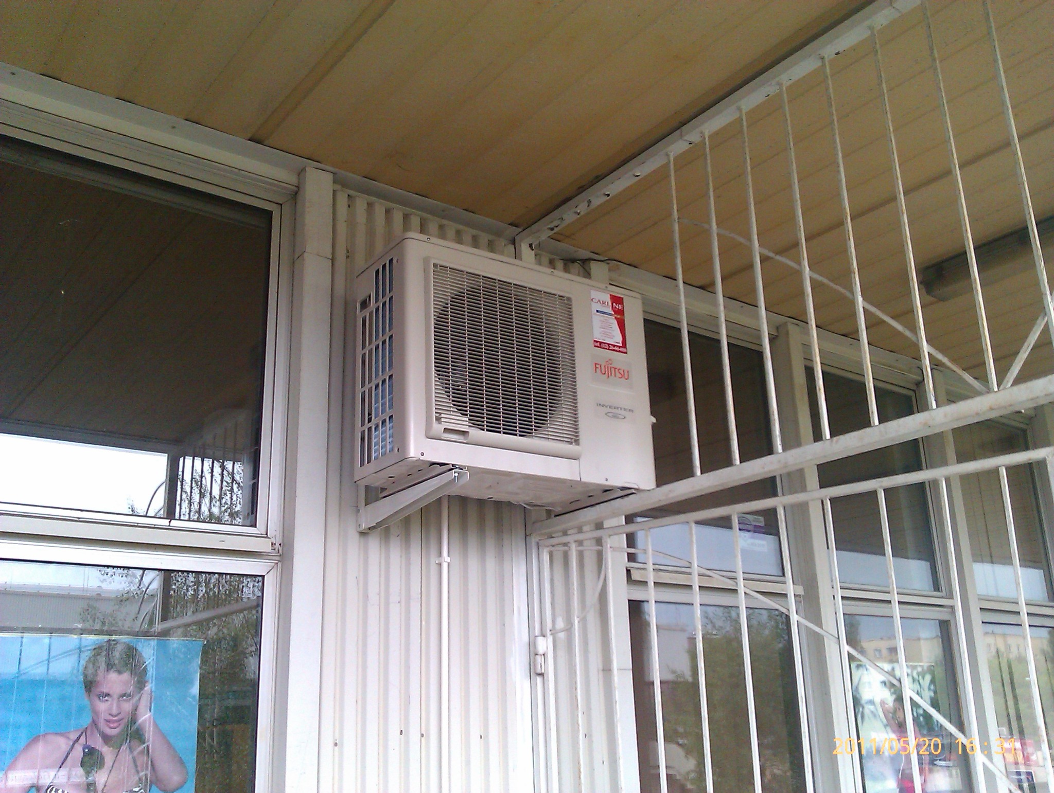 Zewnętrzna jednostka klimatyzacji Fujitsu zamontowana na wspornikach przy ścianie budynku, widoczna przez kratę z białych rurek, z reklamą w oknie w tle.