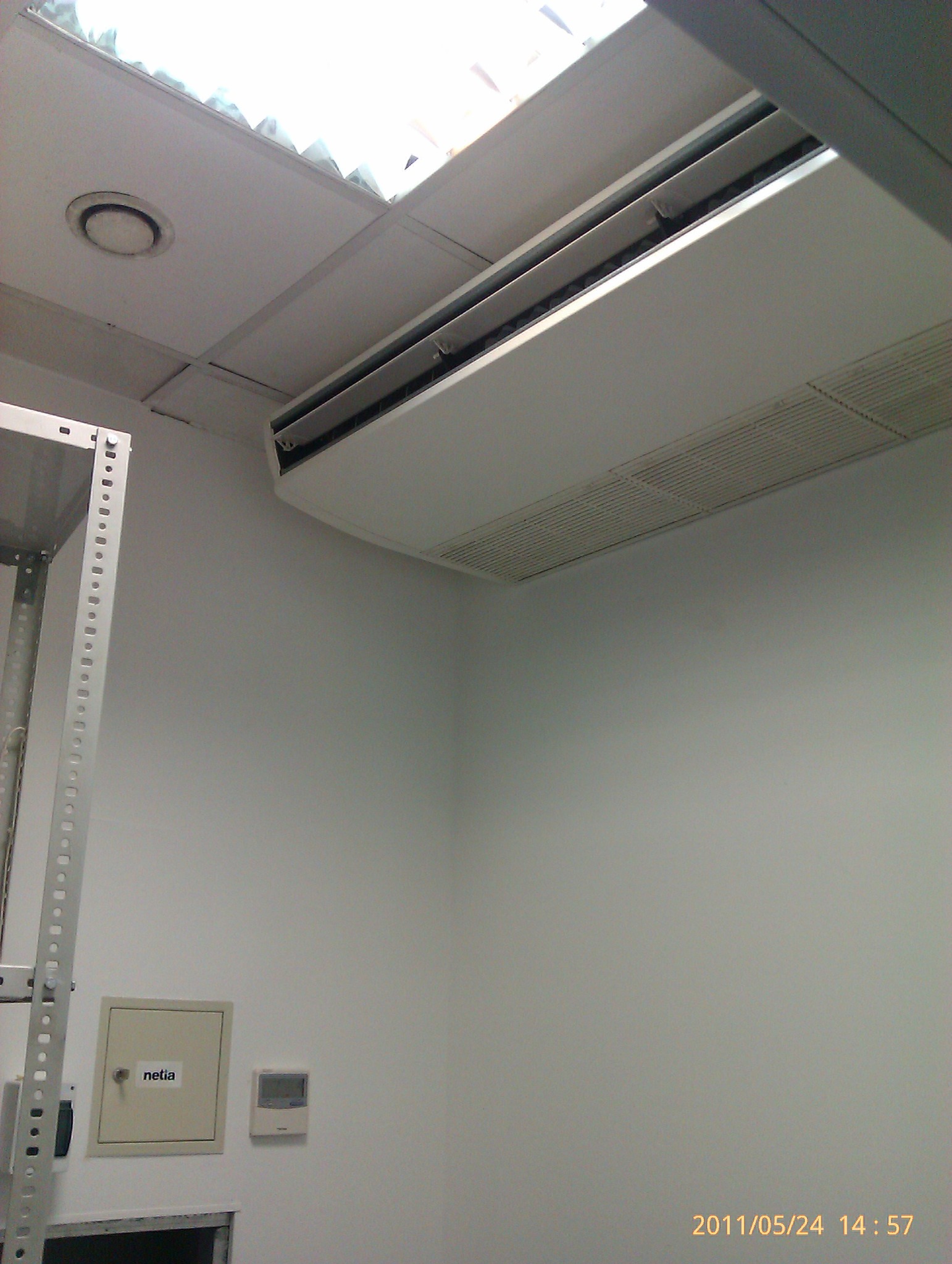 Sufitowa jednostka klimatyzacyjna w biurze, widoczna kratka wentylacyjna i fragment metalowego regału, na ścianie puszka elektryczna z naklejką 'netia' i panel sterowania.