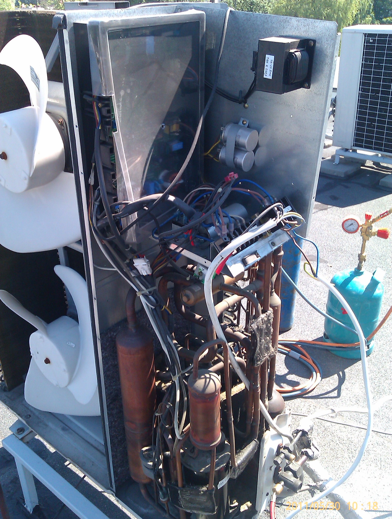 Otwarta obudowa zewnętrznej jednostki klimatyzacji z widocznymi elementami wewnętrznymi: wentylator, rurki miedziane, okablowanie oraz butla z gazem i manometrem na dachu.