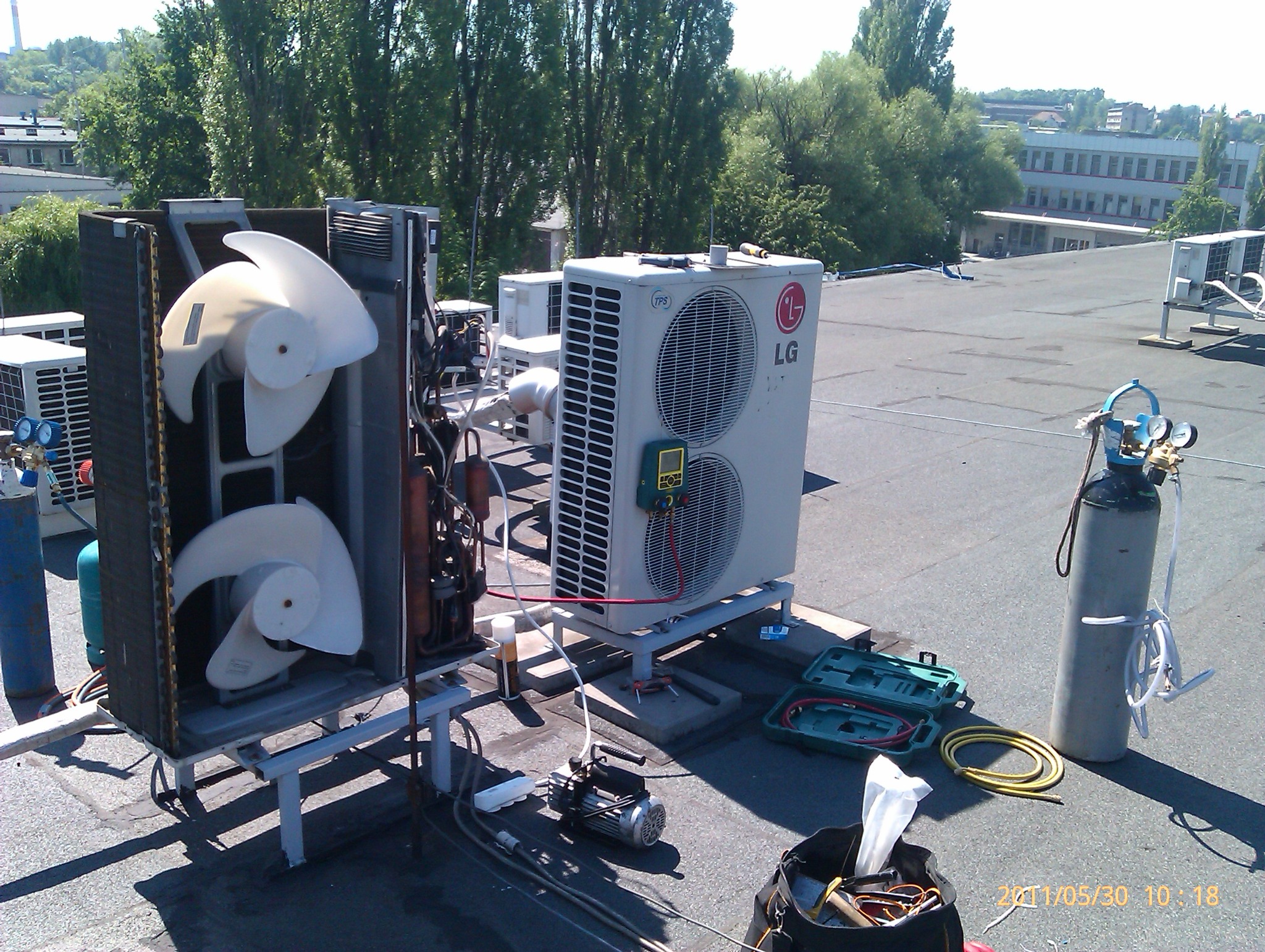 Montaż klimatyzatorów LG na dachu budynku, widoczne narzędzia i butle z gazem, wentylatory oraz miernik podłączony do urządzenia.