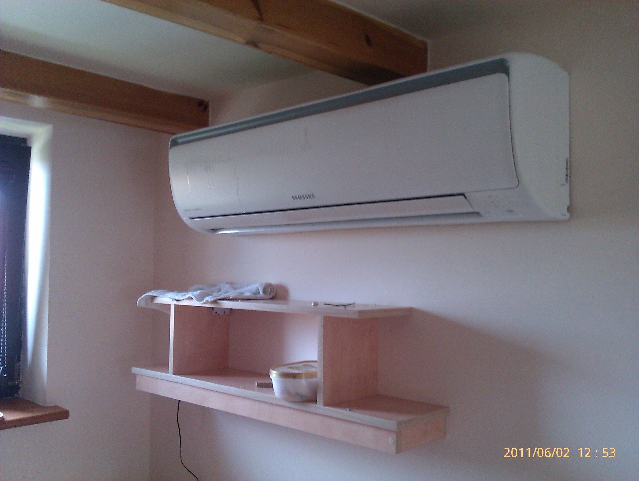 Zamontowany klimatyzator Samsung na ścianie w pomieszczeniu w Krakowie, widoczna drewniana półka pod spodem i fragment okna.