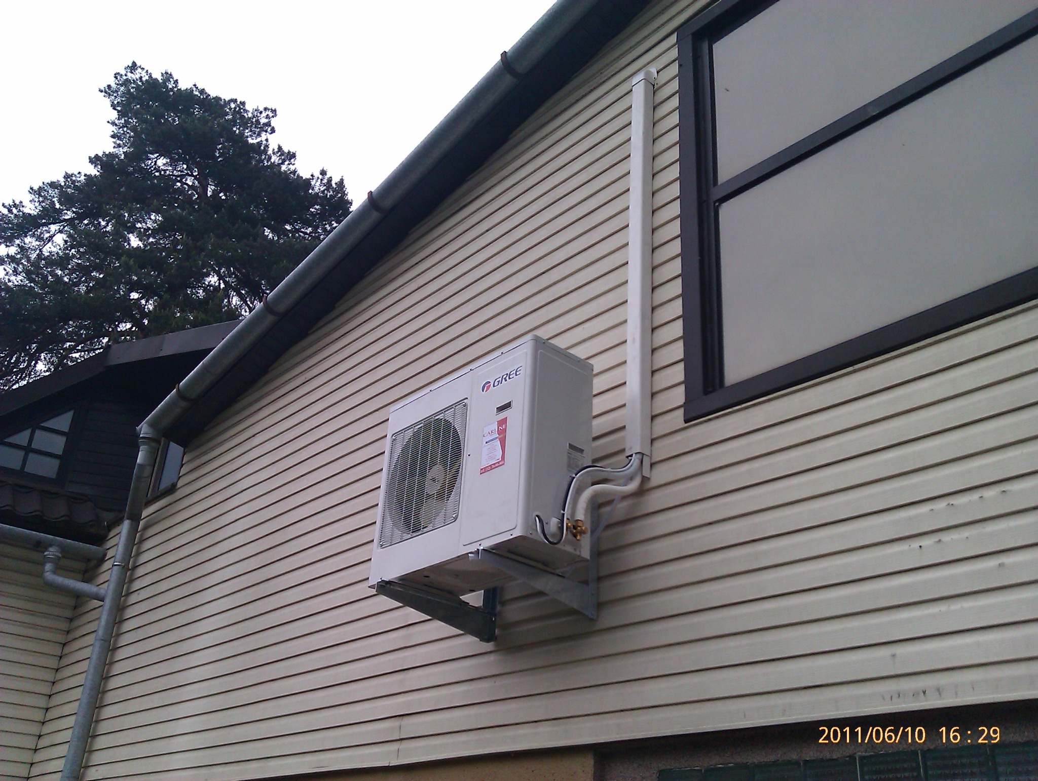 Zewnętrzna jednostka klimatyzacji marki Gree zamontowana na ścianie budynku z sidingiem, widoczne rury i elementy montażowe.
