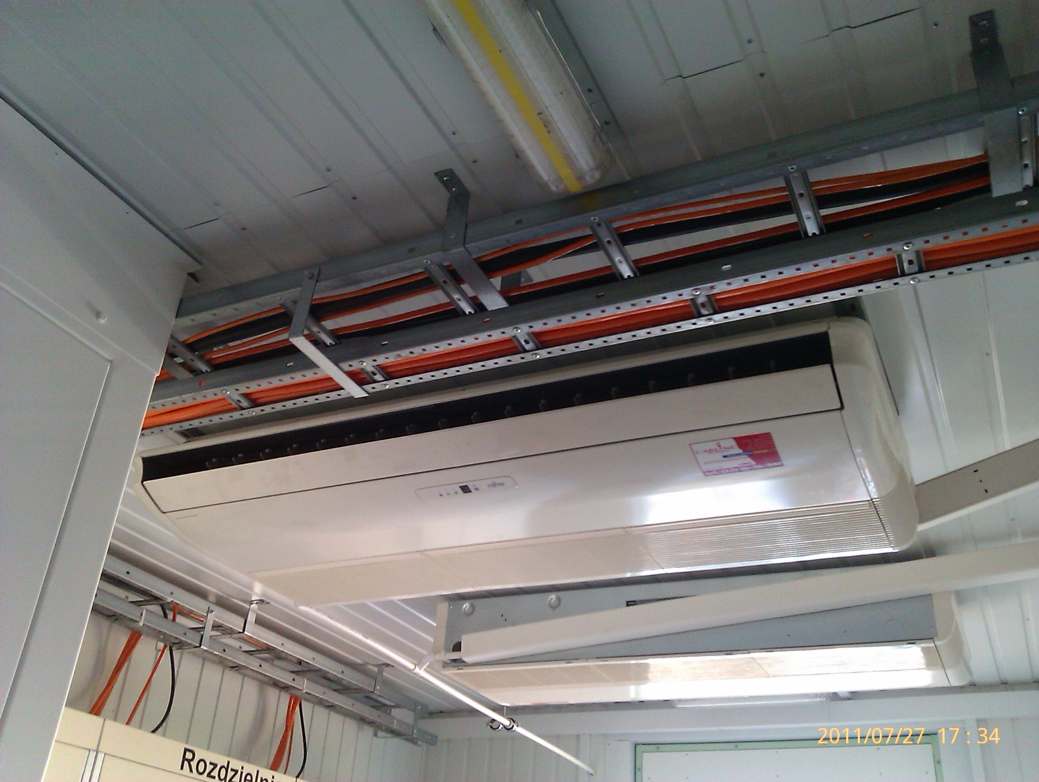 Klimatyzator zamontowany pod sufitem w pomieszczeniu z widocznymi kablami i metalowymi konstrukcjami wsporczymi.