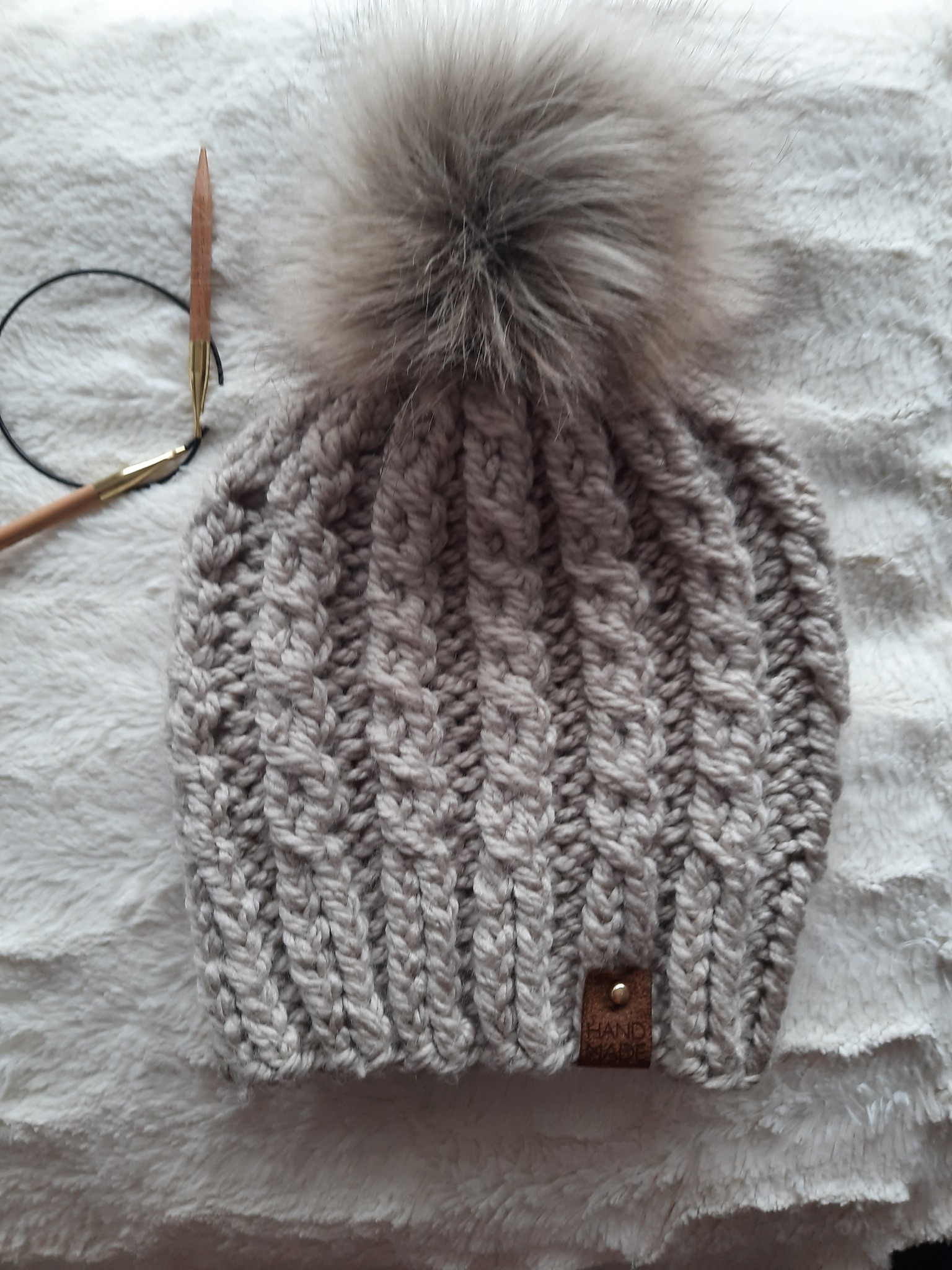 Ręcznie robiona, szara czapka z pomponem z futra, leżąca na puszystym, białym tle, obok drewniane druty dziewiarskie z metalowymi końcówkami.