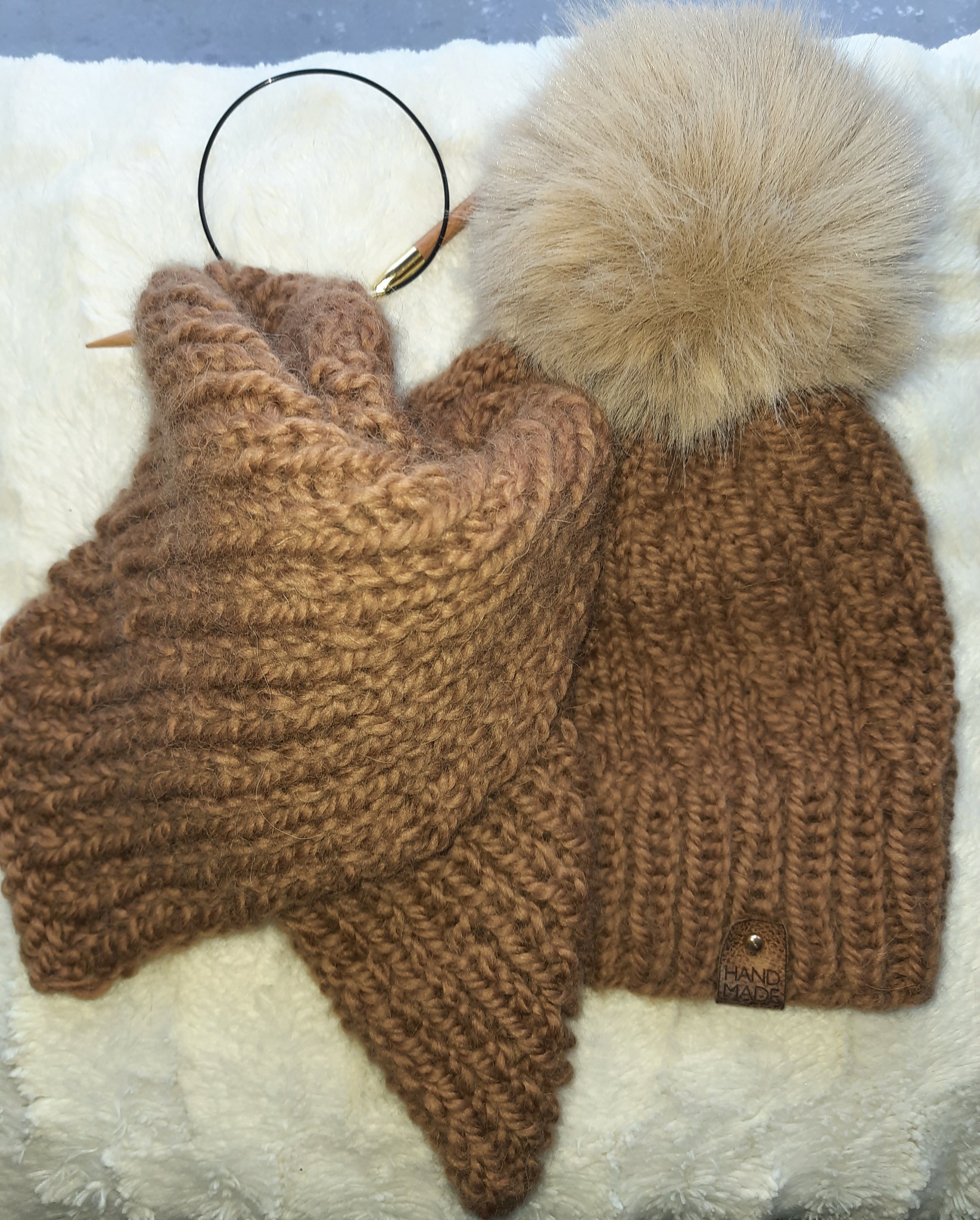 Brązowy, ręcznie robiony komplet: czapka z pomponem i szalik, leżące na białym, puszystym tle, obok druty dziewiarskie.