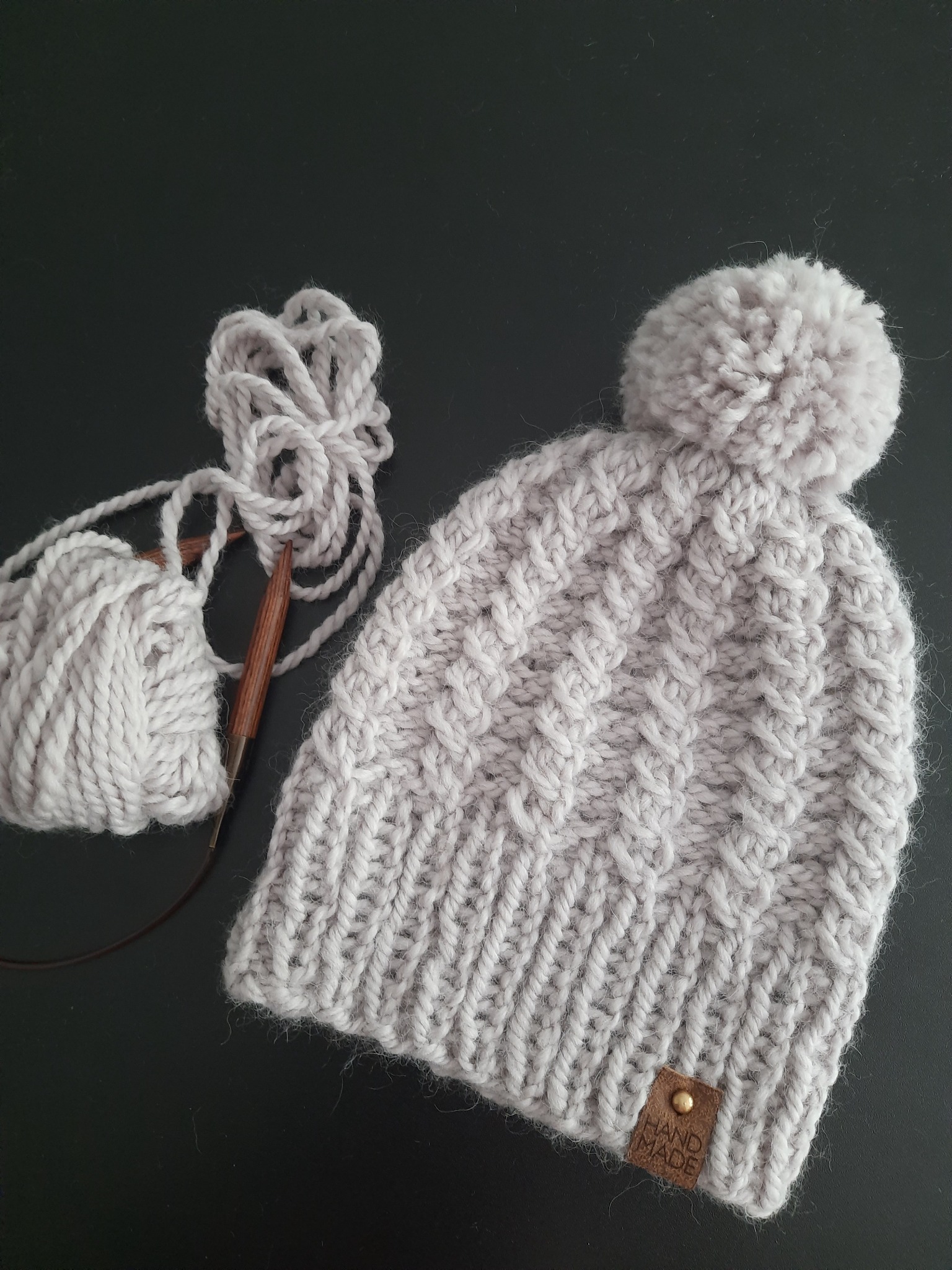 Ręcznie robiona, szara czapka z pomponem, leżąca obok kłębka włóczki i drewnianych drutów dziewiarskich na ciemnym tle, z widoczną metką 'Hand Made'.