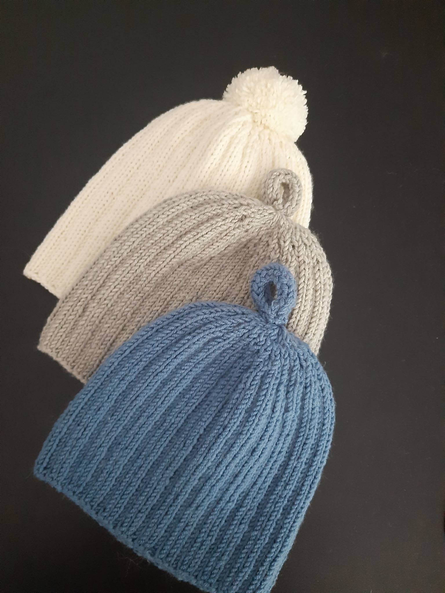Trzy ręcznie robione, dziergane czapki w odcieniach bieli, szarości i błękitu, ułożone jedna za drugą na czarnym tle. Biała czapka z pomponem na górze.