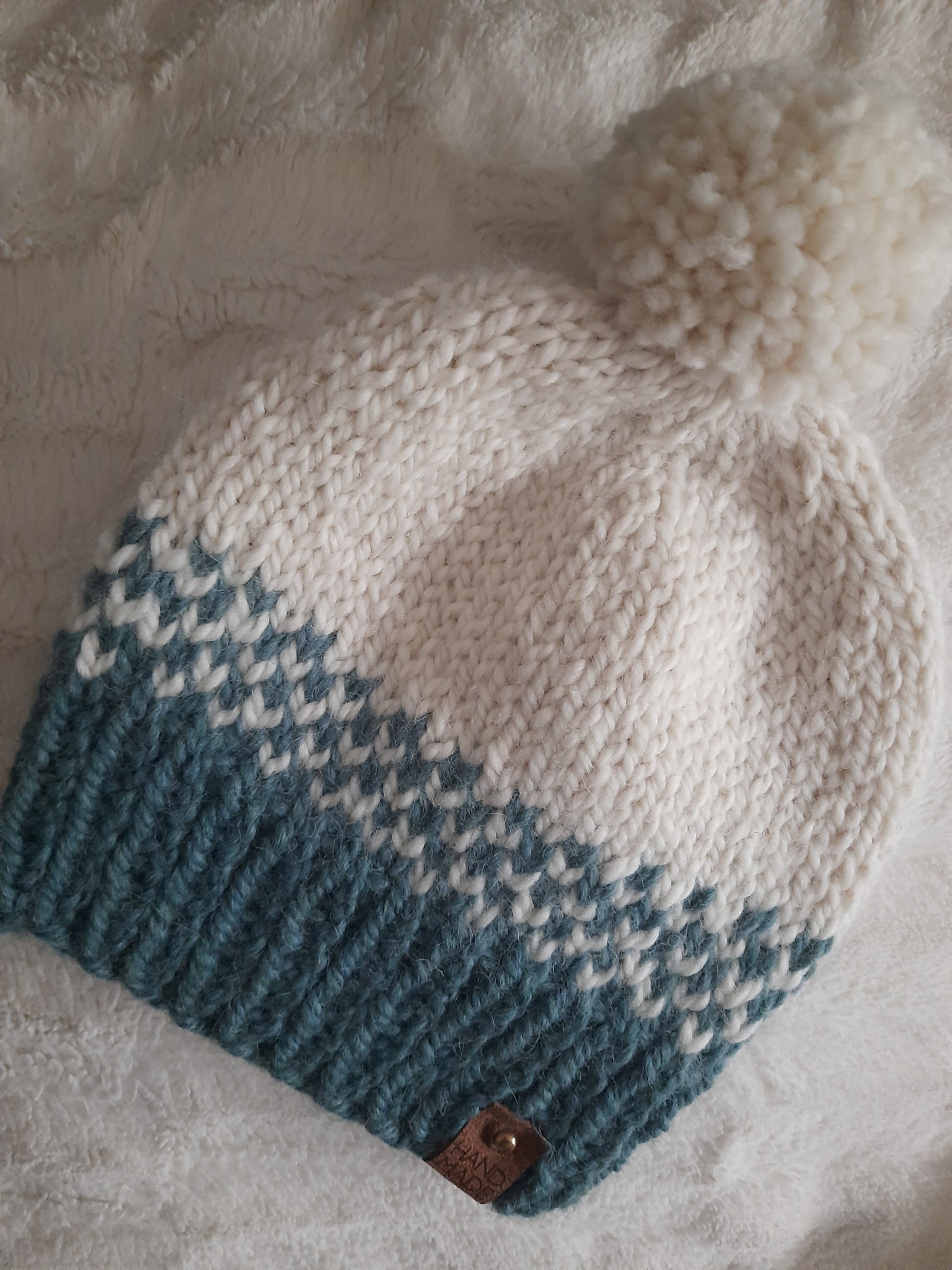 Ręcznie robiona, dziergana czapka z pomponem, w kolorze białym i turkusowym, leżąca na miękkim, białym tle, z widoczną skórzaną metką z logo.