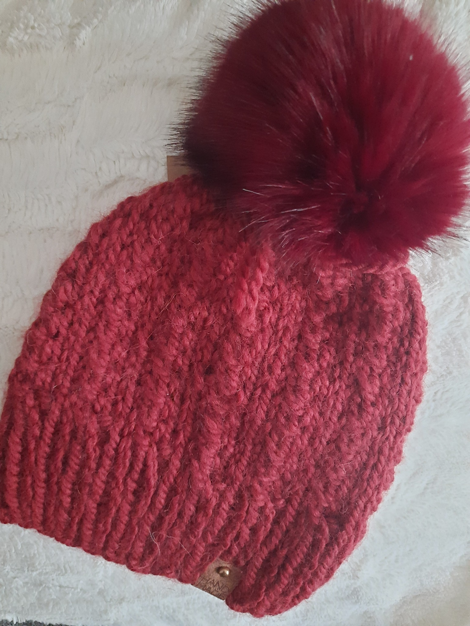 Czerwona, ręcznie robiona czapka z grubym splotem i dużym, puszystym pomponem na białym, puszystym tle.