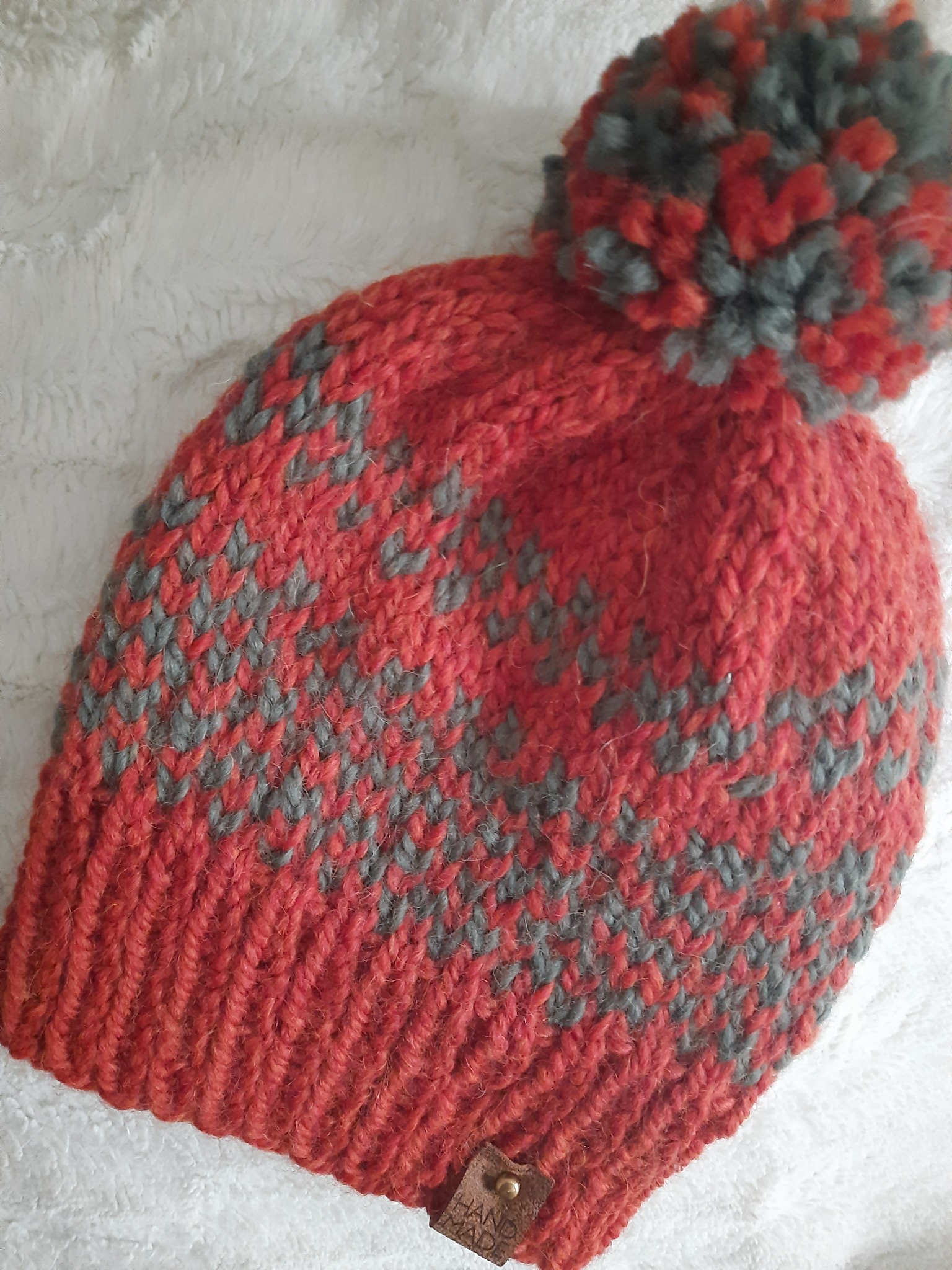 Pomarańczowo-szara czapka z pomponem, wykonana na drutach, leżąca na białym puszystym tle, z małą skórzaną metką 'Hand Made'.