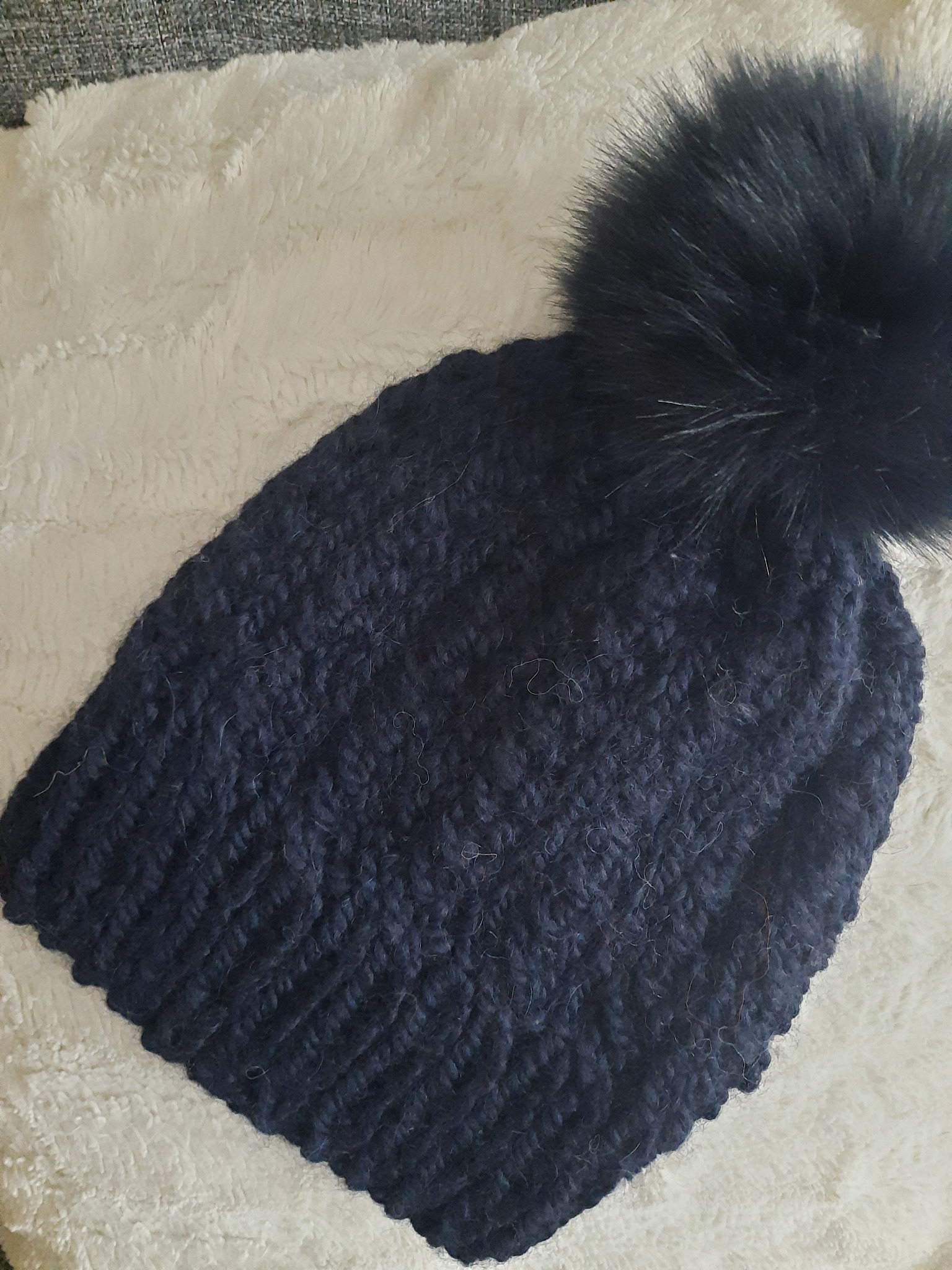 Ciemnoniebieska, ręcznie robiona czapka z grubym splotem i dużym, puszystym pomponem z imitacji futra, leżąca na miękkim, kremowym kocu.