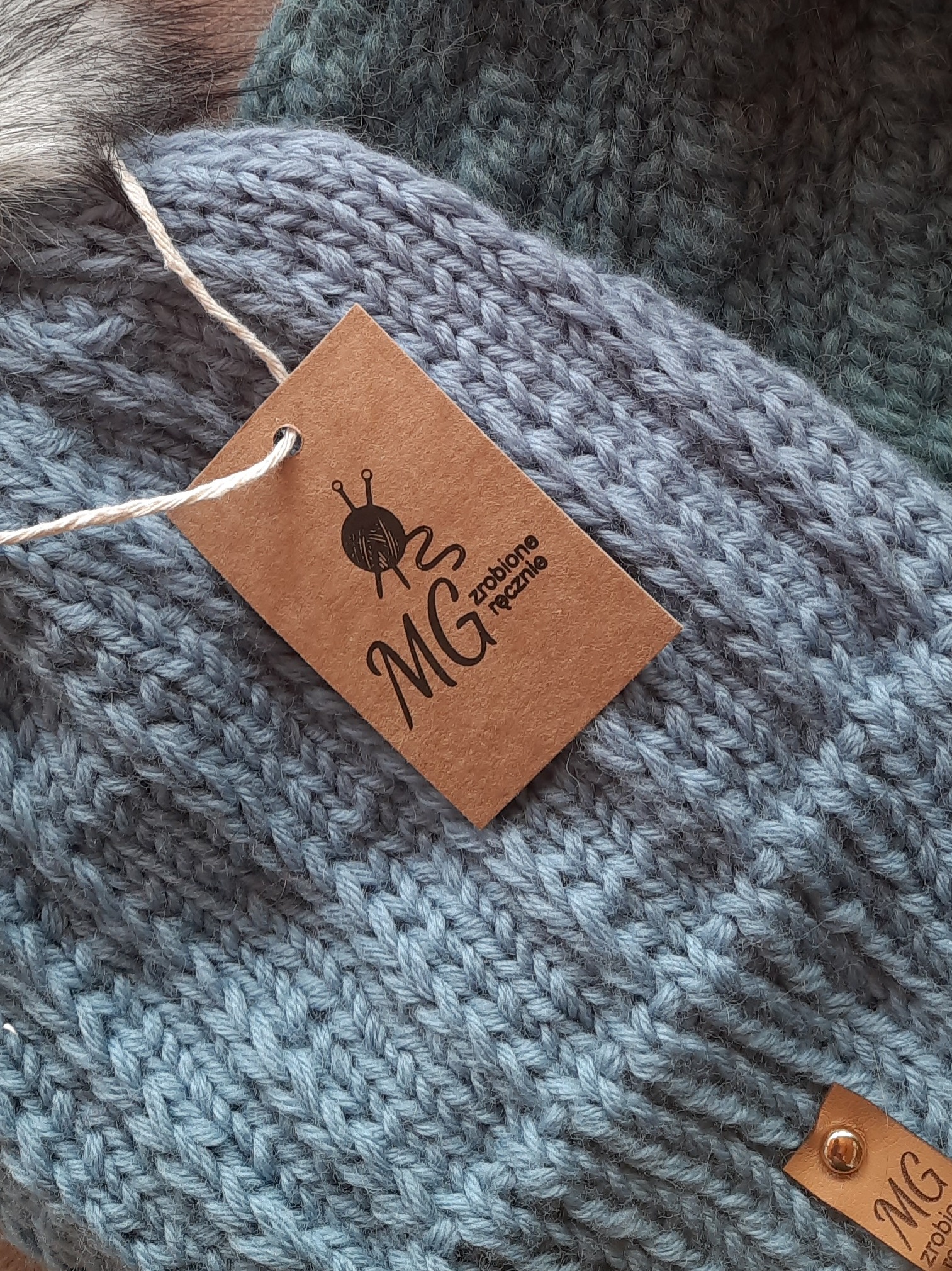 Detale dwóch ręcznie robionych, wełnianych czapek w odcieniach szarości i zieleni, z brązową metką z logo dziewiarskim i napisem 'MG zrobione ręcznie'.