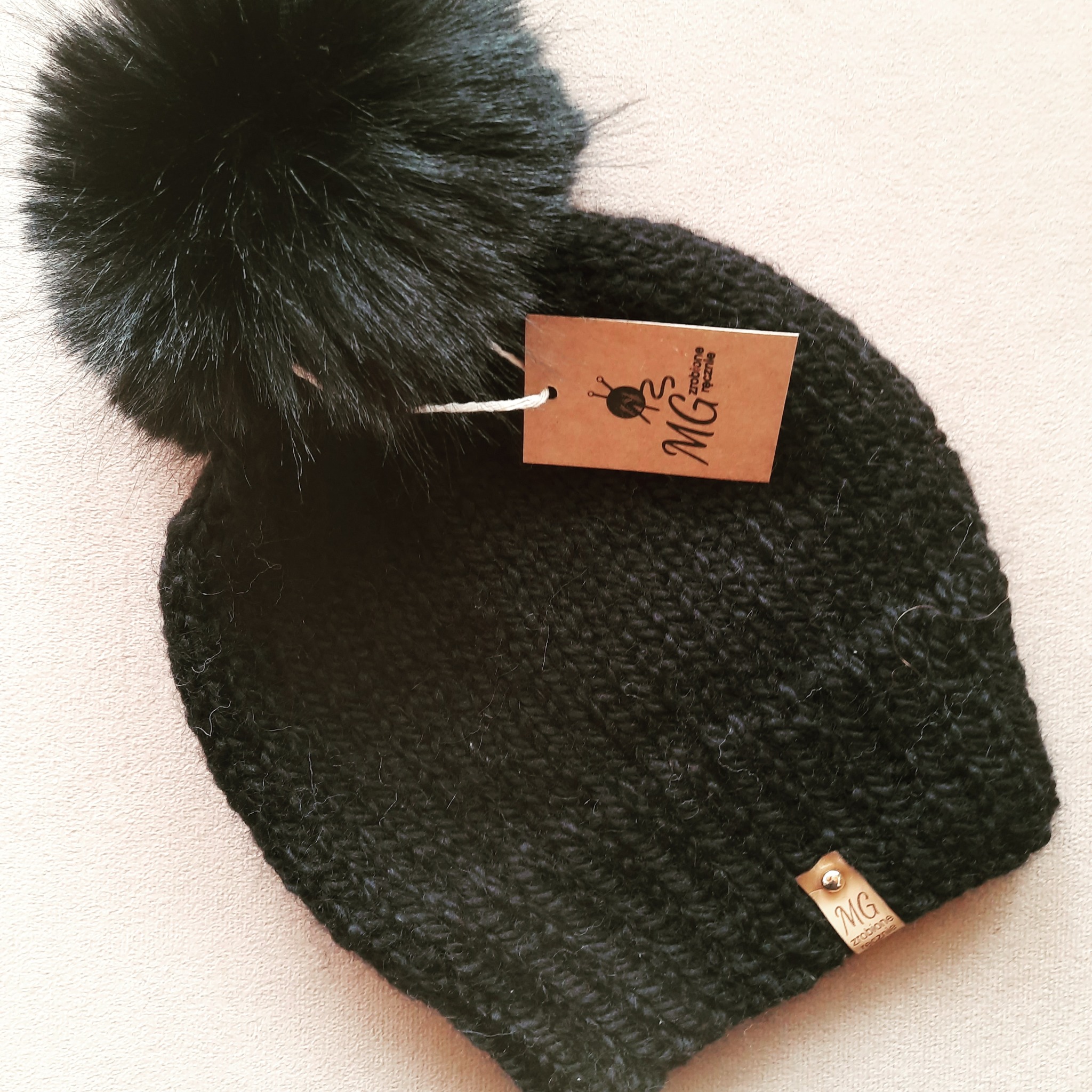 Czarna, ręcznie robiona czapka z pomponem, z metką z logo producenta, leżąca na beżowym tle.