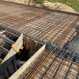 USŁUGI OGÓLNOBUDOWLANE KAMIL JÓSKOWSKI - Zbrojenie fundamentów z częściowo wylanym betonem, widoczne drewniane szalunki i siatka zbrojeniowa. Ujęcie z góry, w tle ziemia i trawa.