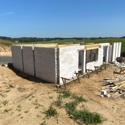 USŁUGI OGÓLNOBUDOWLANE KAMIL JÓSKOWSKI - Budowa domu w toku: ściany z betonu komórkowego, zbrojenie, drewniane podpory. W tle zielone pola i błękitne niebo. Widok z poziomu gruntu, w słoneczny dzień.