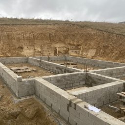 USŁUGI OGÓLNOBUDOWLANE KAMIL JÓSKOWSKI - Wysoki kąt na fundamenty z bloczków betonowych, zbrojenie pionowe, wykop ziemny w tle. Biała szmatka leży na murze. Pochmurne niebo.